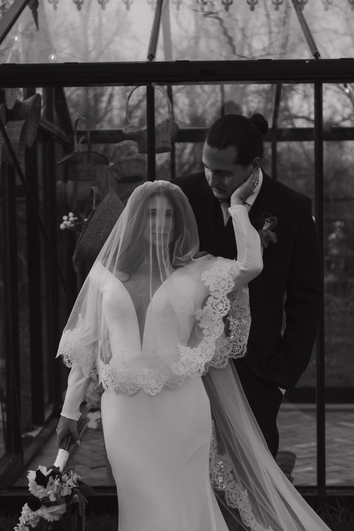 Black and white portrait of bride in lace veil and gown with groom outside glass greenhouse.