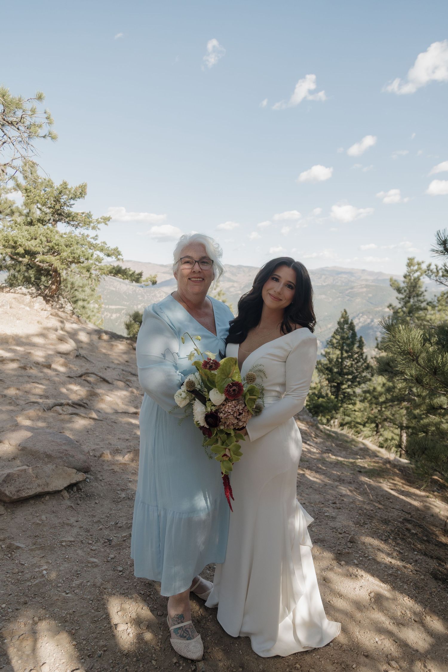 Bride in white gown holds bouquet while posing with older woman in blue dress on mountain overlook.