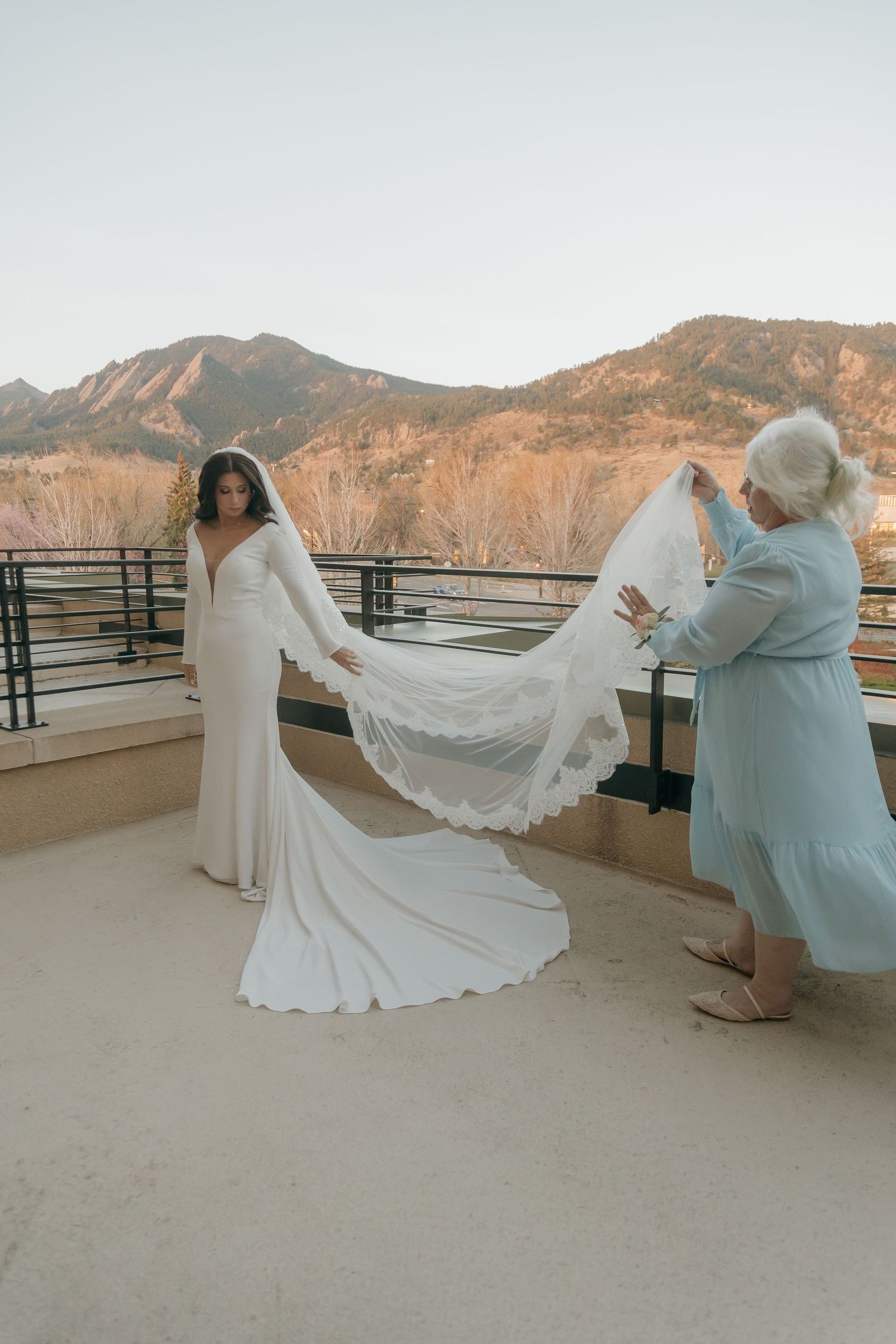 Bride in white gown and veil on mountain terrace while woman adjusts her train with Flatirons backdrop.