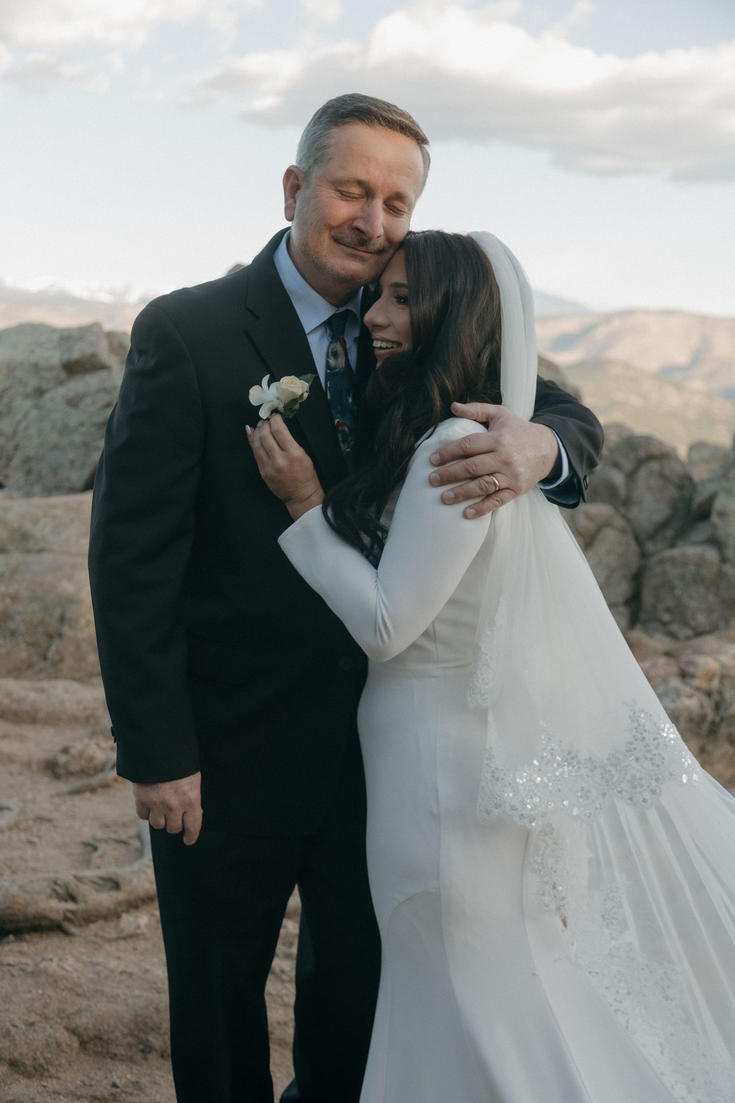 Bride in white gown and veil embracing man in black suit at rocky mountain overlook.