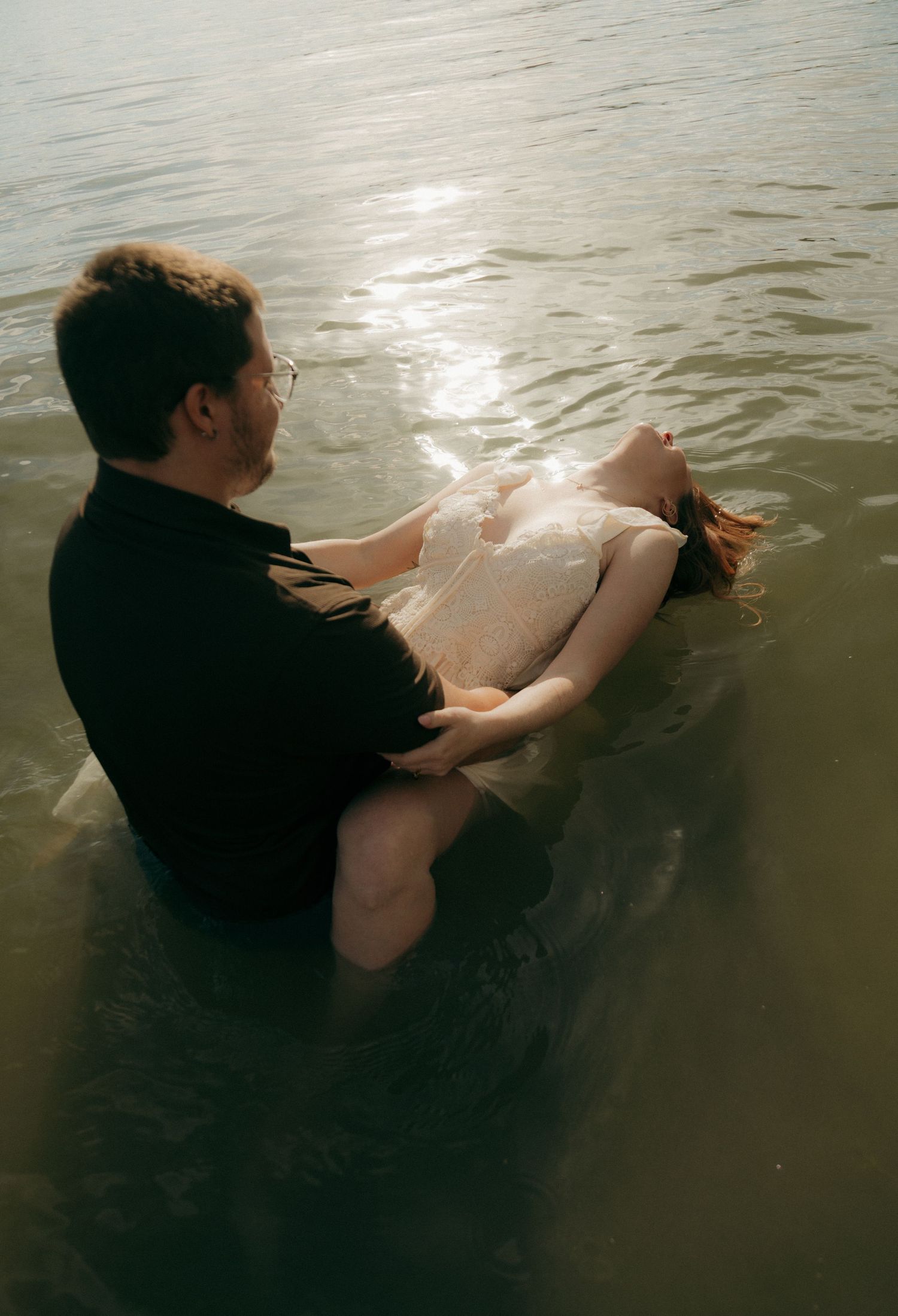 Man holds woman in white dress while wading in sunlit water during romantic couples photo session.