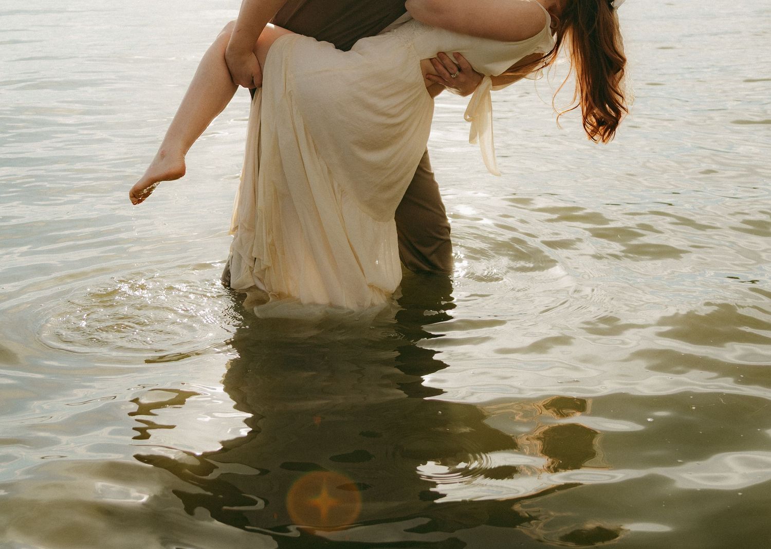 Person carrying a woman in a white dress while wading through shallow water outdoors.