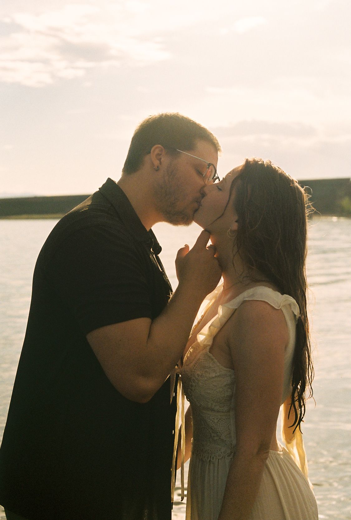 Couple sharing a tender kiss backlit by warm sunset glow near a waterfront.