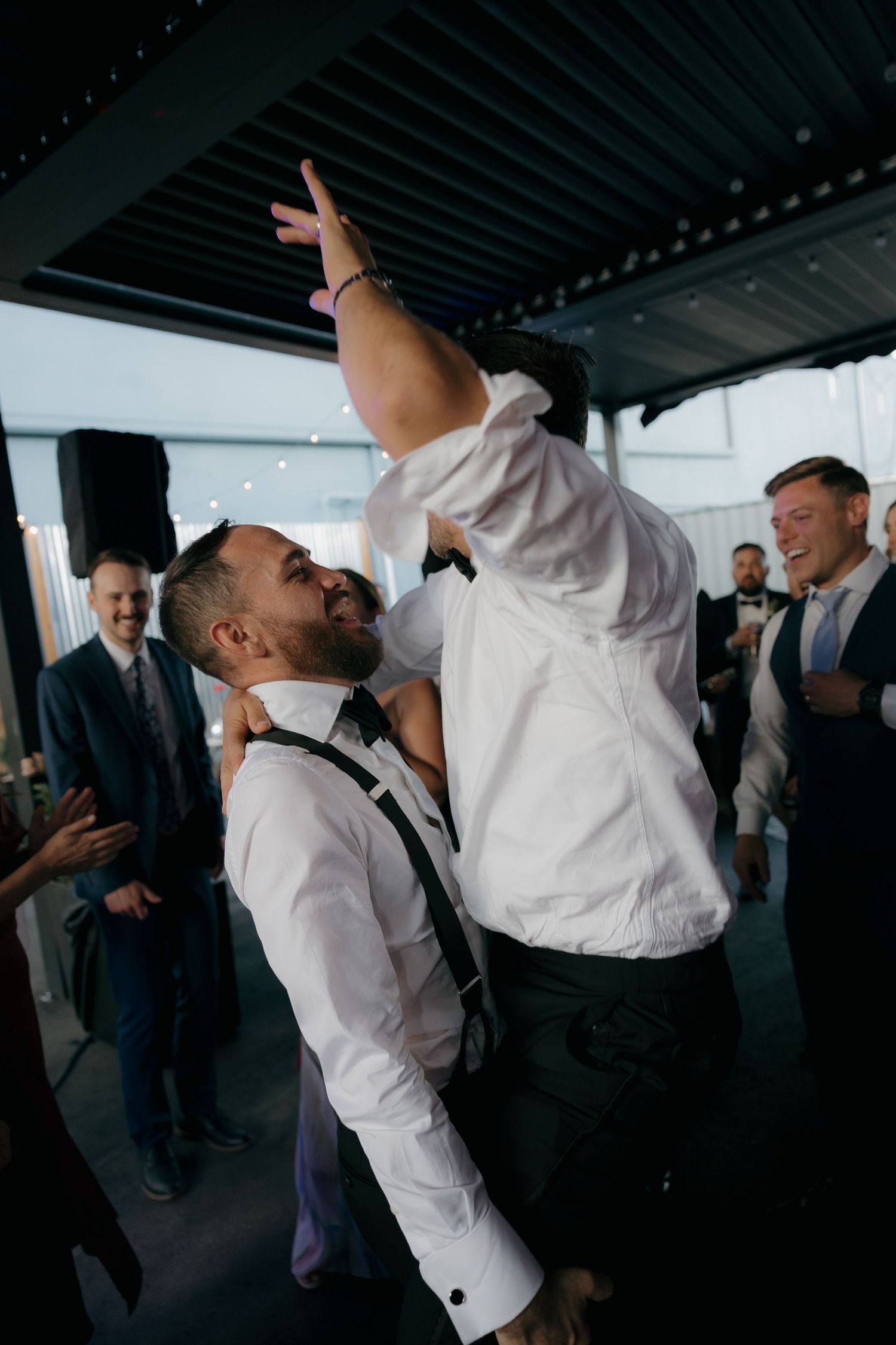 Energetic wedding reception dance floor moment with groomsmen celebrating in white dress shirts and suspenders.