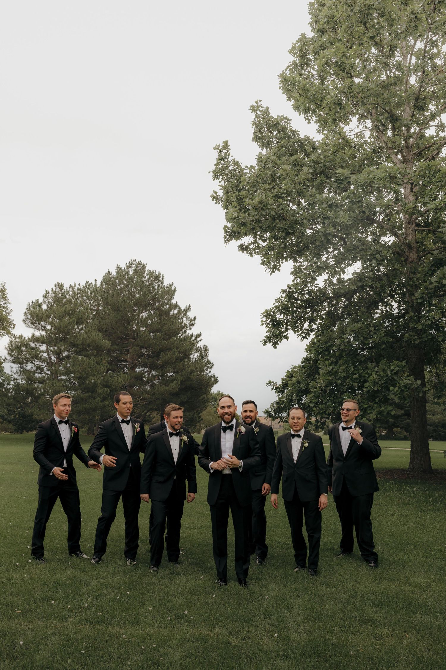 Groom and groomsmen in black tuxedos walking together on green lawn outdoors.