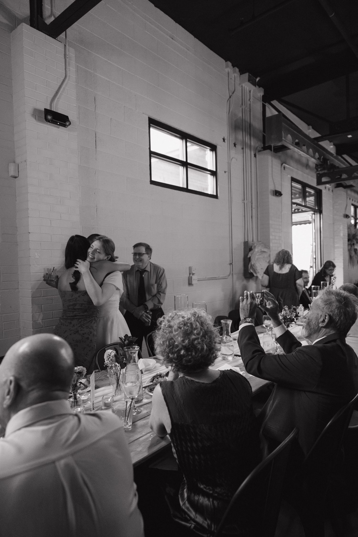 Black and white photo of wedding guests celebrating at reception dinner in industrial venue.