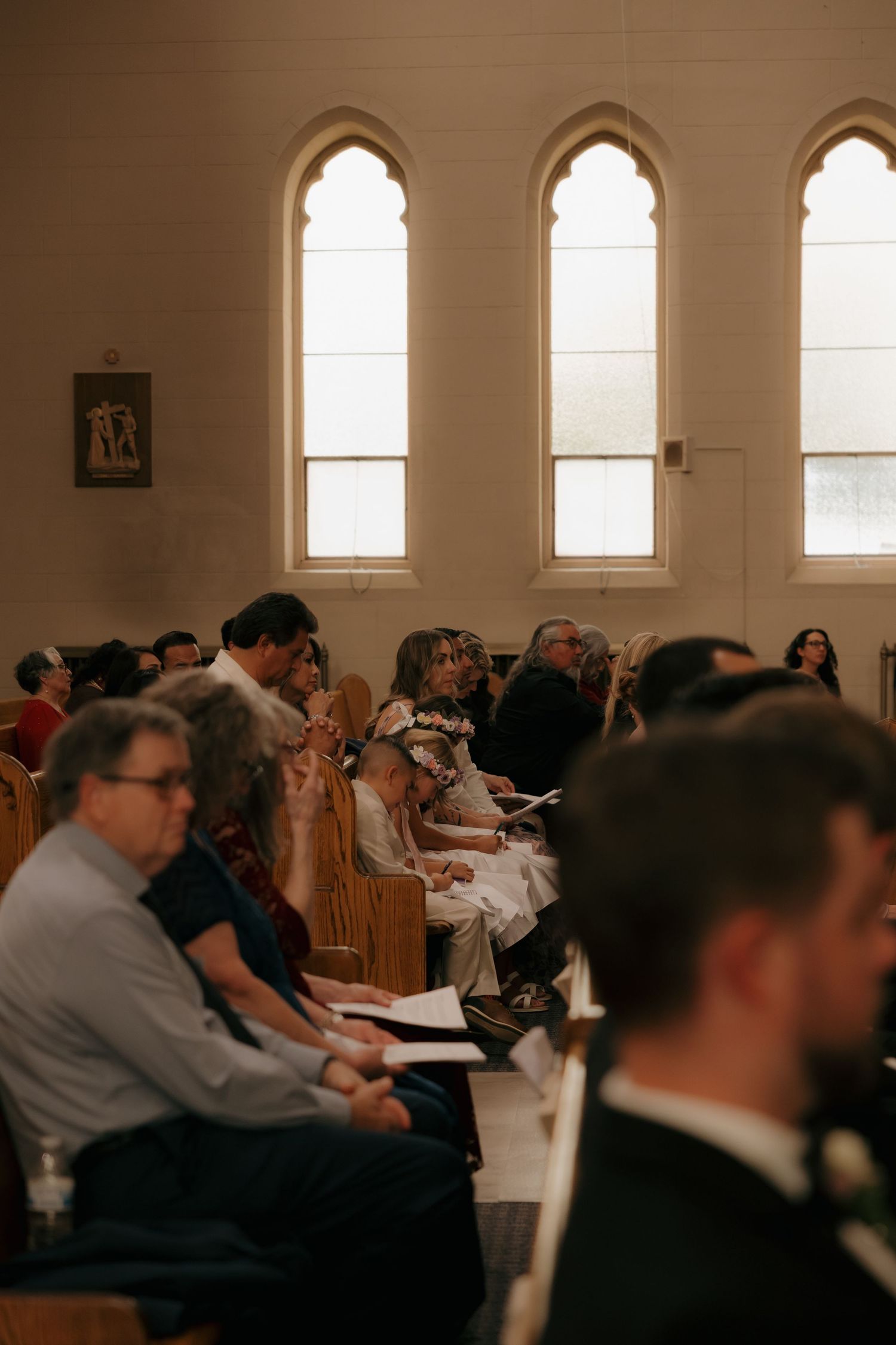 Wedding guests seated in church pews during a ceremony, with tall arched windows letting in soft natural light.