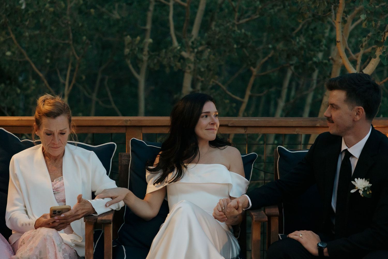 Bride in strapless white gown smiles while seated between two guests at outdoor wedding reception.