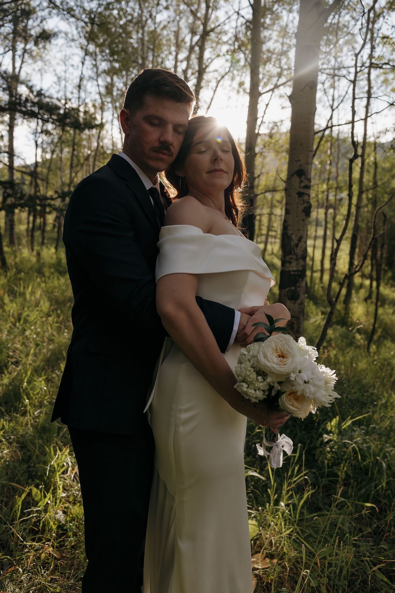 Groom embraces bride from behind in sunlit forest as she holds white peony bouquet.