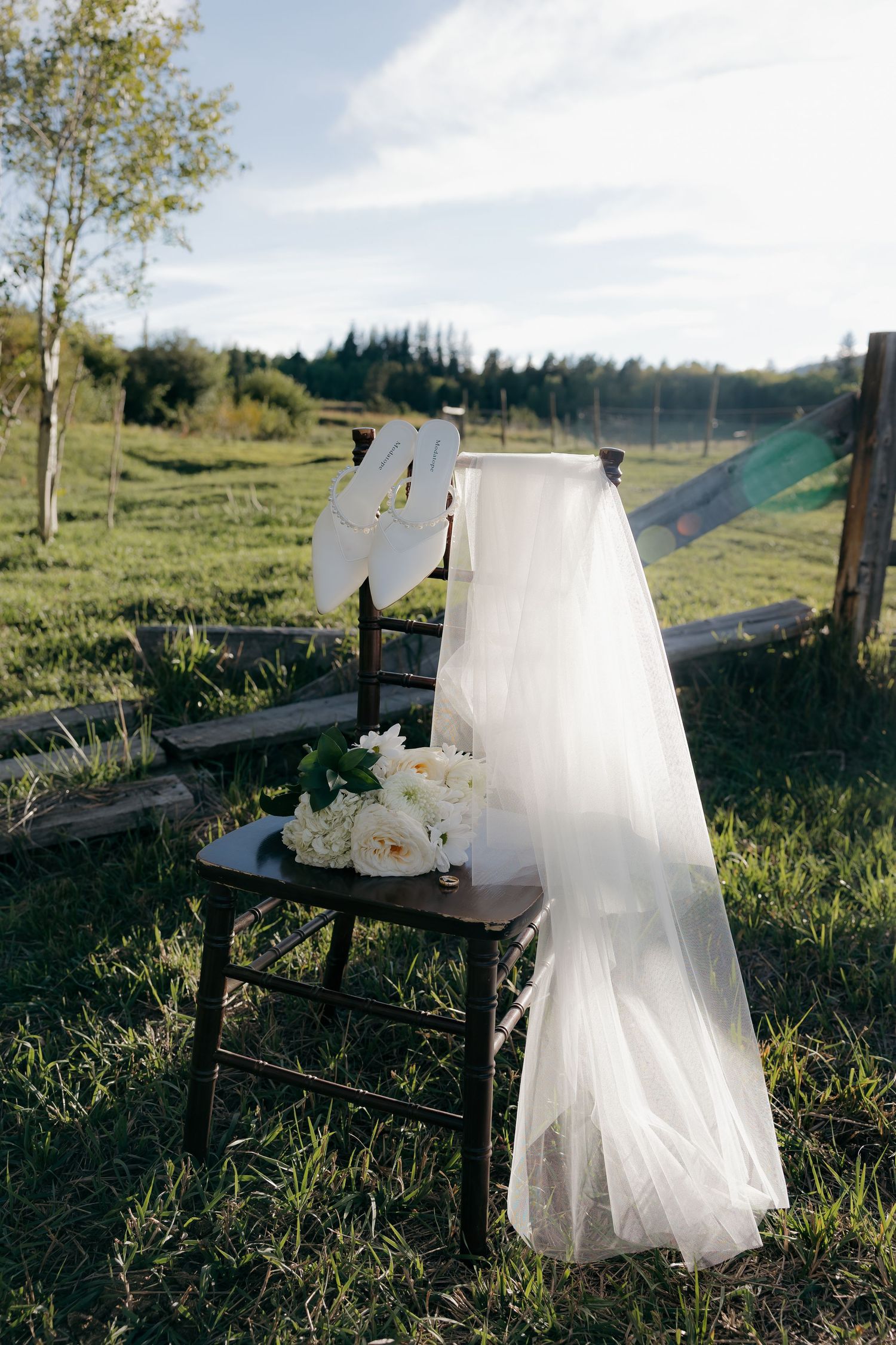 Elegant white wedding veil and bridal bouquet of white roses draped over a rustic wooden chair in a green field.