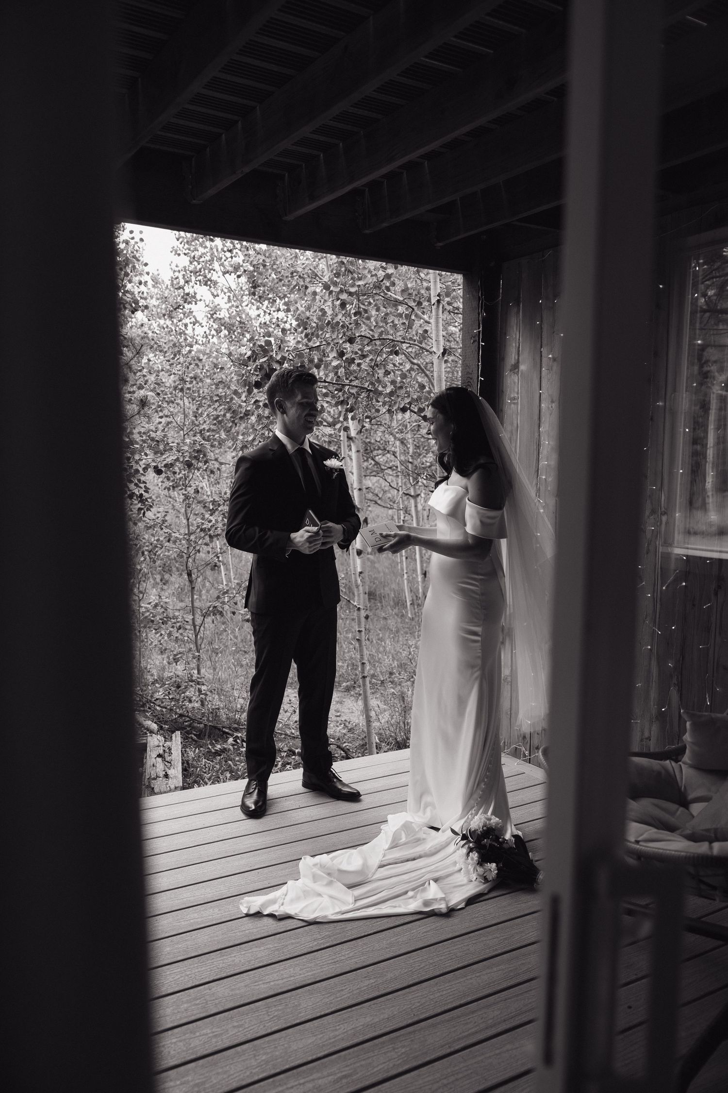 Black and white photo of bride and groom exchanging vows on a wooden deck surrounded by trees.