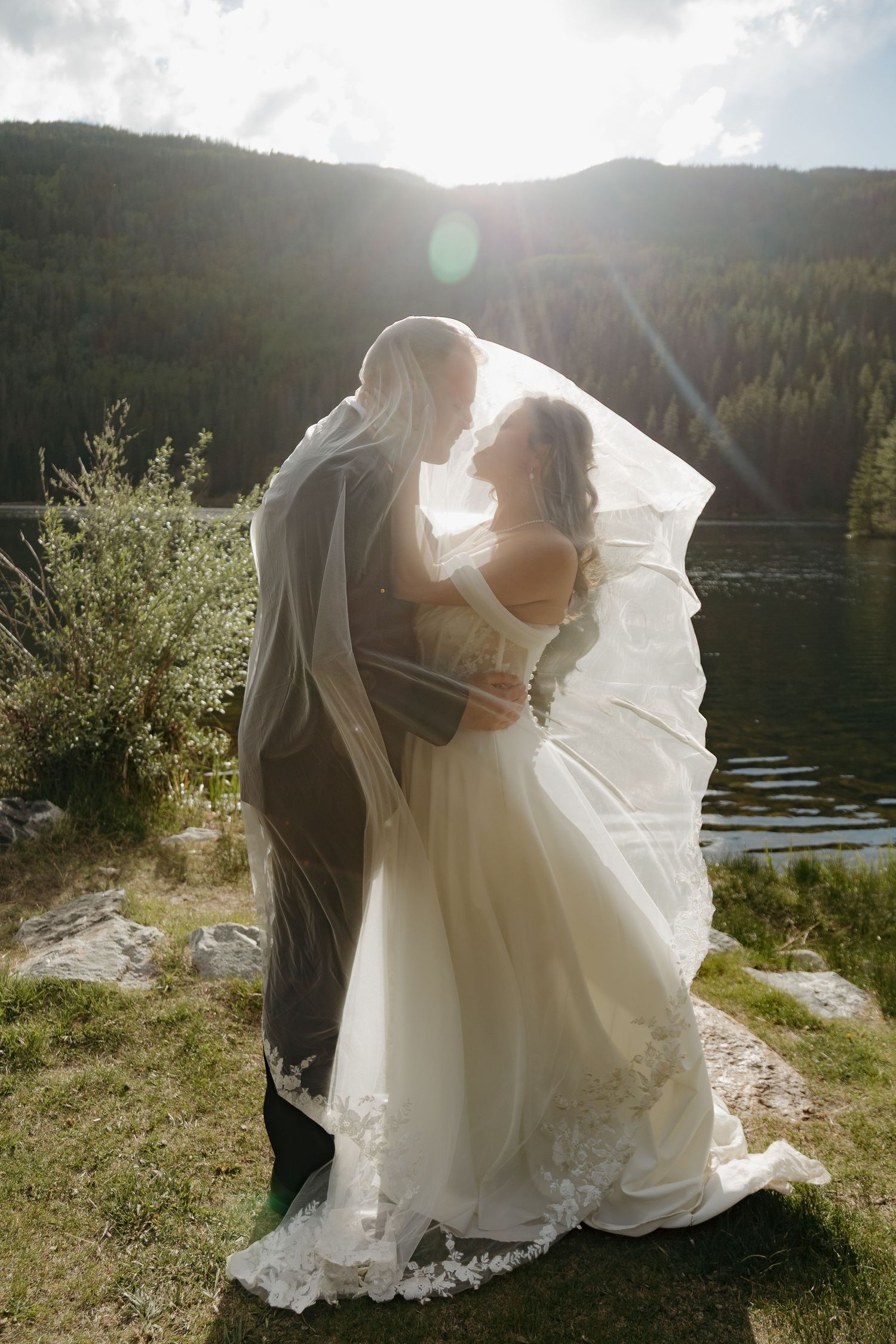 Bride and groom share romantic moment under flowing veil by mountain lake with sunlight streaming behind them.