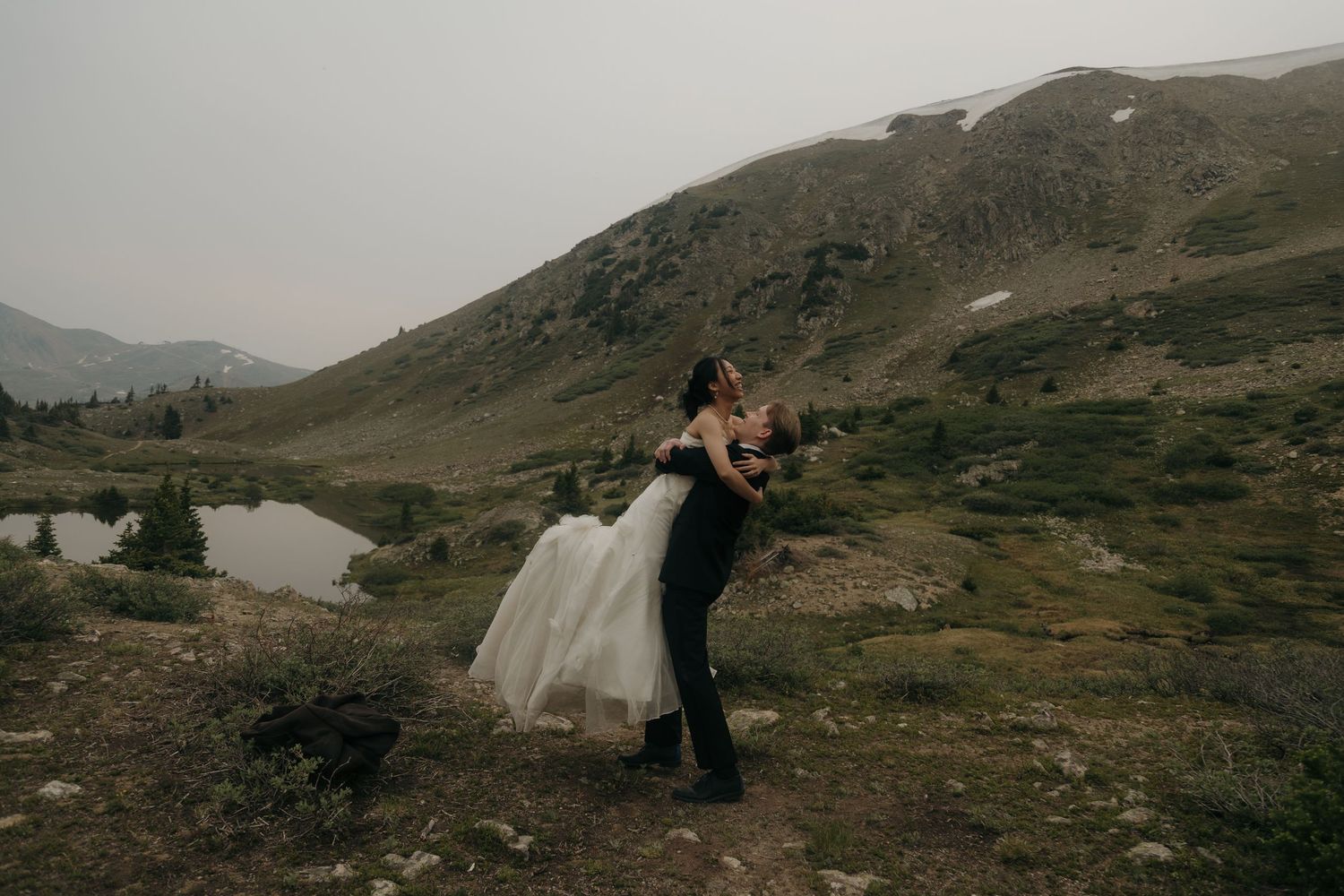 Groom lifting bride in wedding attire with mountain lake backdrop during outdoor elopement session.
