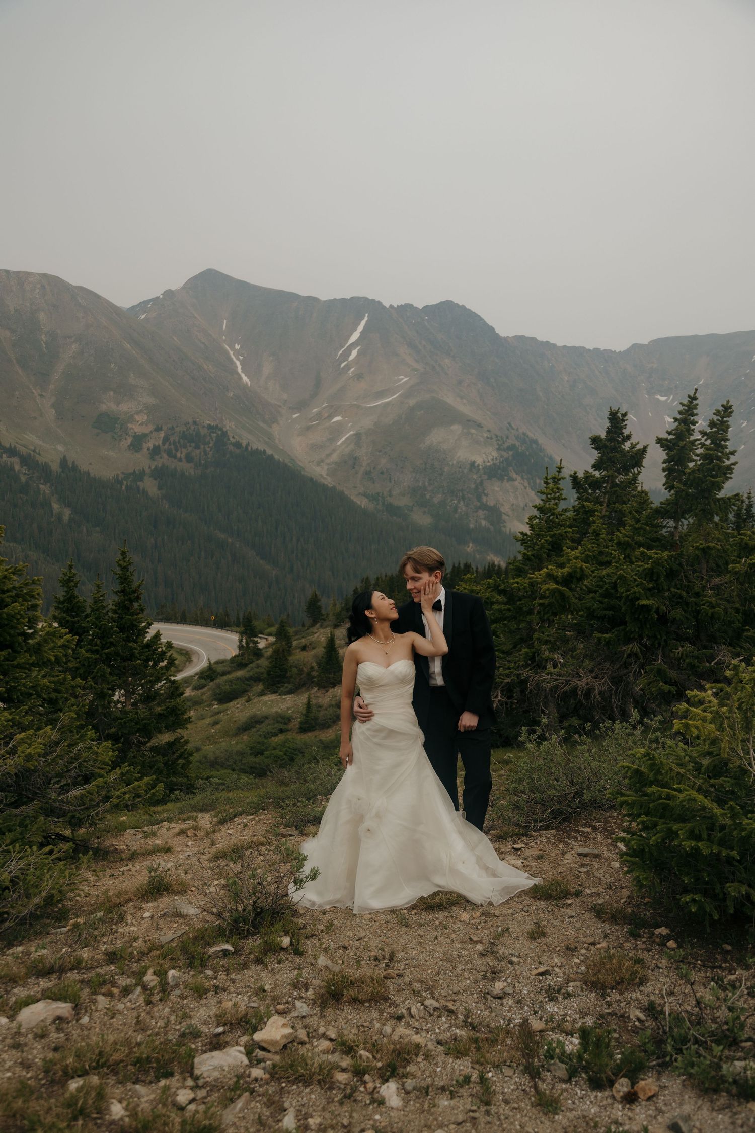 Wedding couple standing together on a rocky mountain trail surrounded by pine trees and misty peaks.