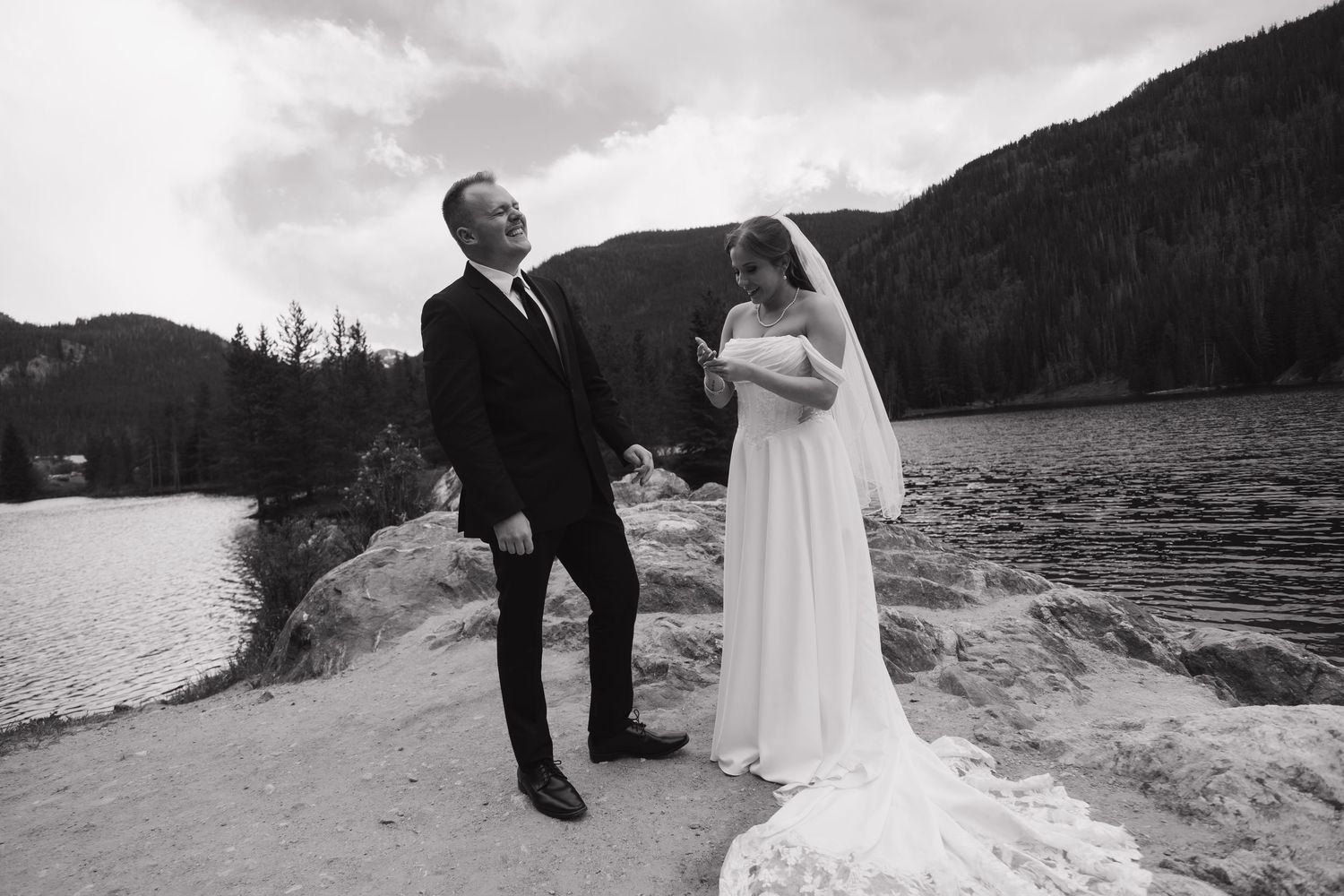 Black and white photo of a bride and groom sharing a joyful moment by a mountain lake on their wedding day.