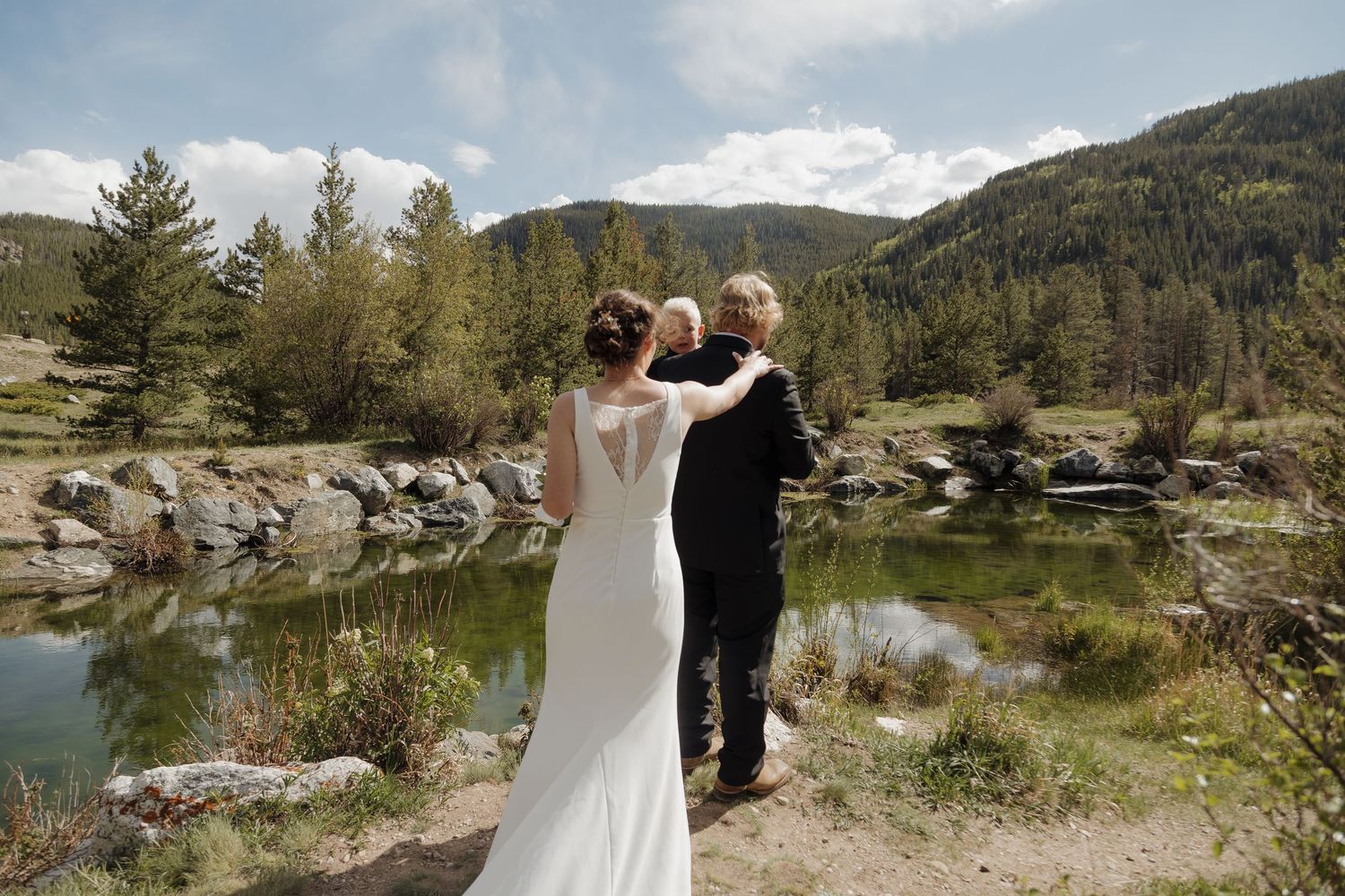 Bride and groom stand together by a mountain pond surrounded by pine trees and rocky landscape on their wedding day.