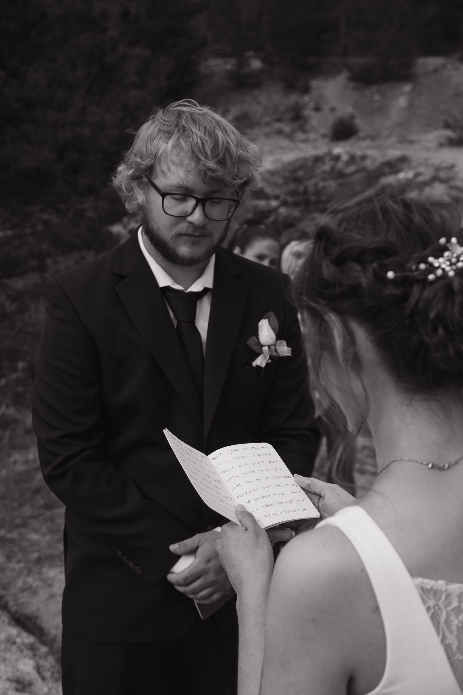 Groom in black suit reads wedding vows from paper during outdoor ceremony.