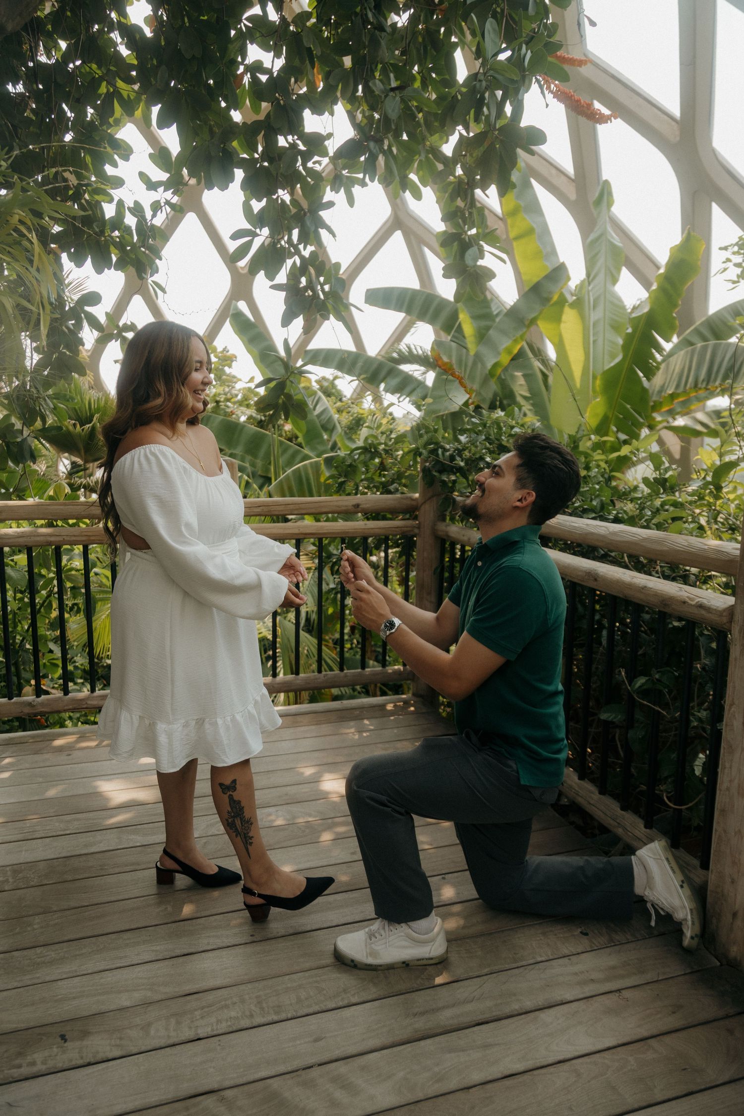 Man kneeling on one knee proposing to woman in white dress on wooden deck in tropical greenhouse.
