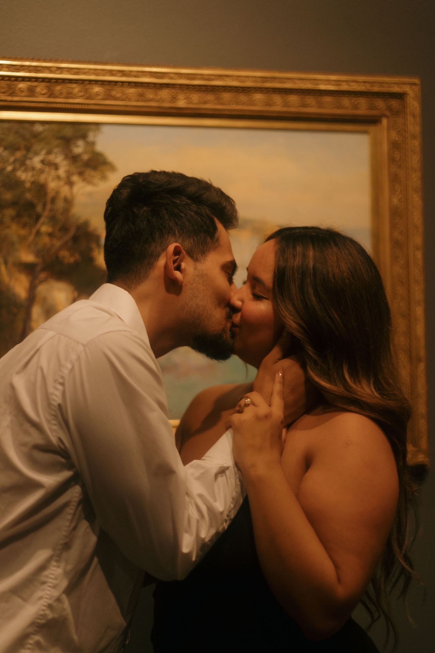 Couple sharing passionate kiss in front of ornate gold-framed painting in dimly lit art gallery.
