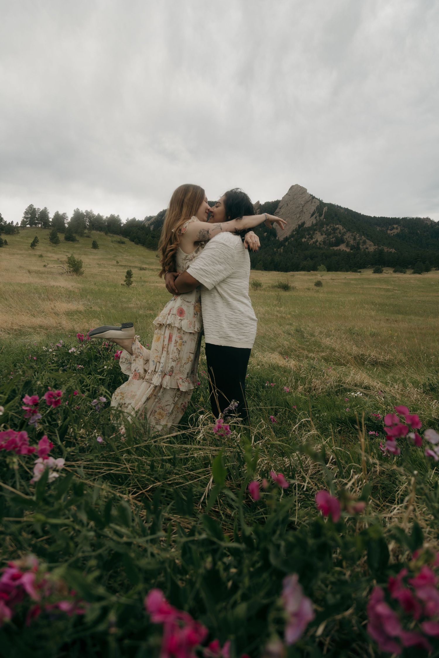 A couple sharing a romantic kiss in a lush green meadow with pink wildflowers and the Flatirons in the background.