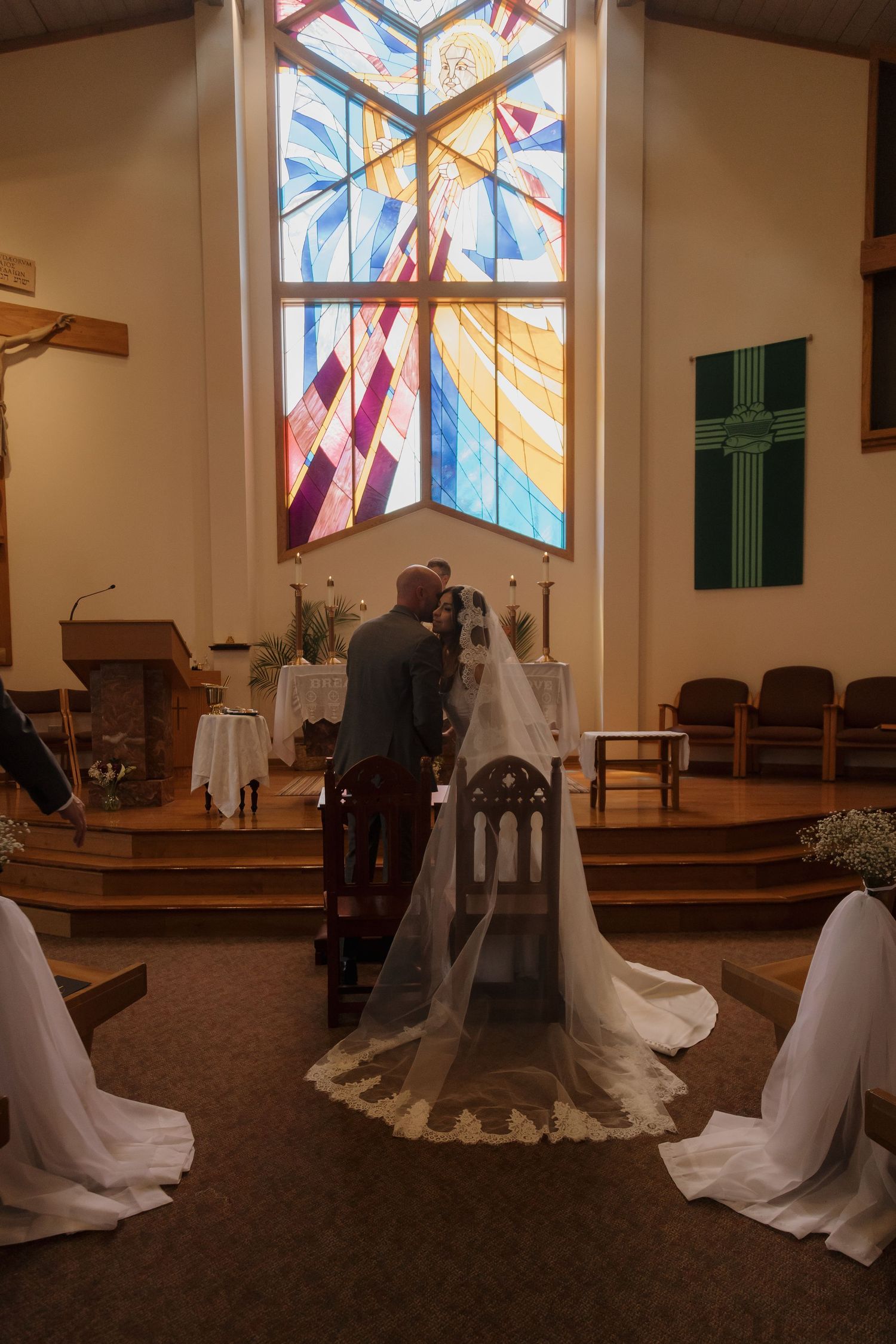 A bride and groom kneel at the altar beneath a colorful stained glass window during their wedding.