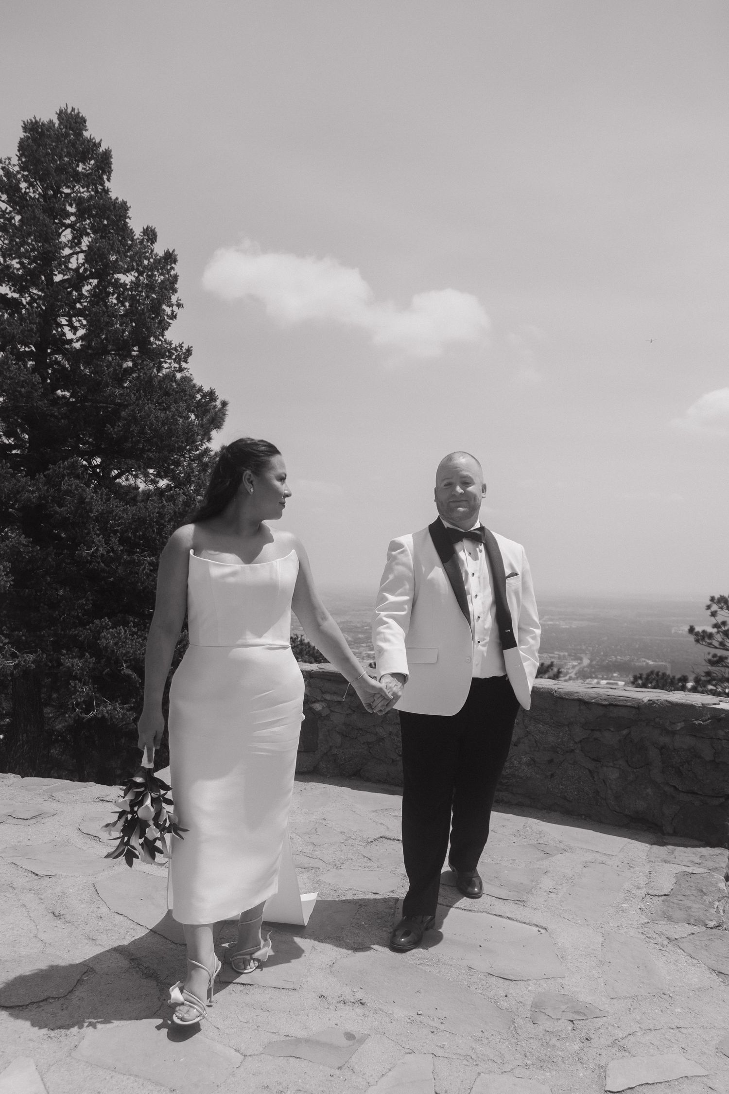 Black and white photo of bride and groom holding hands walking along a stone overlook.