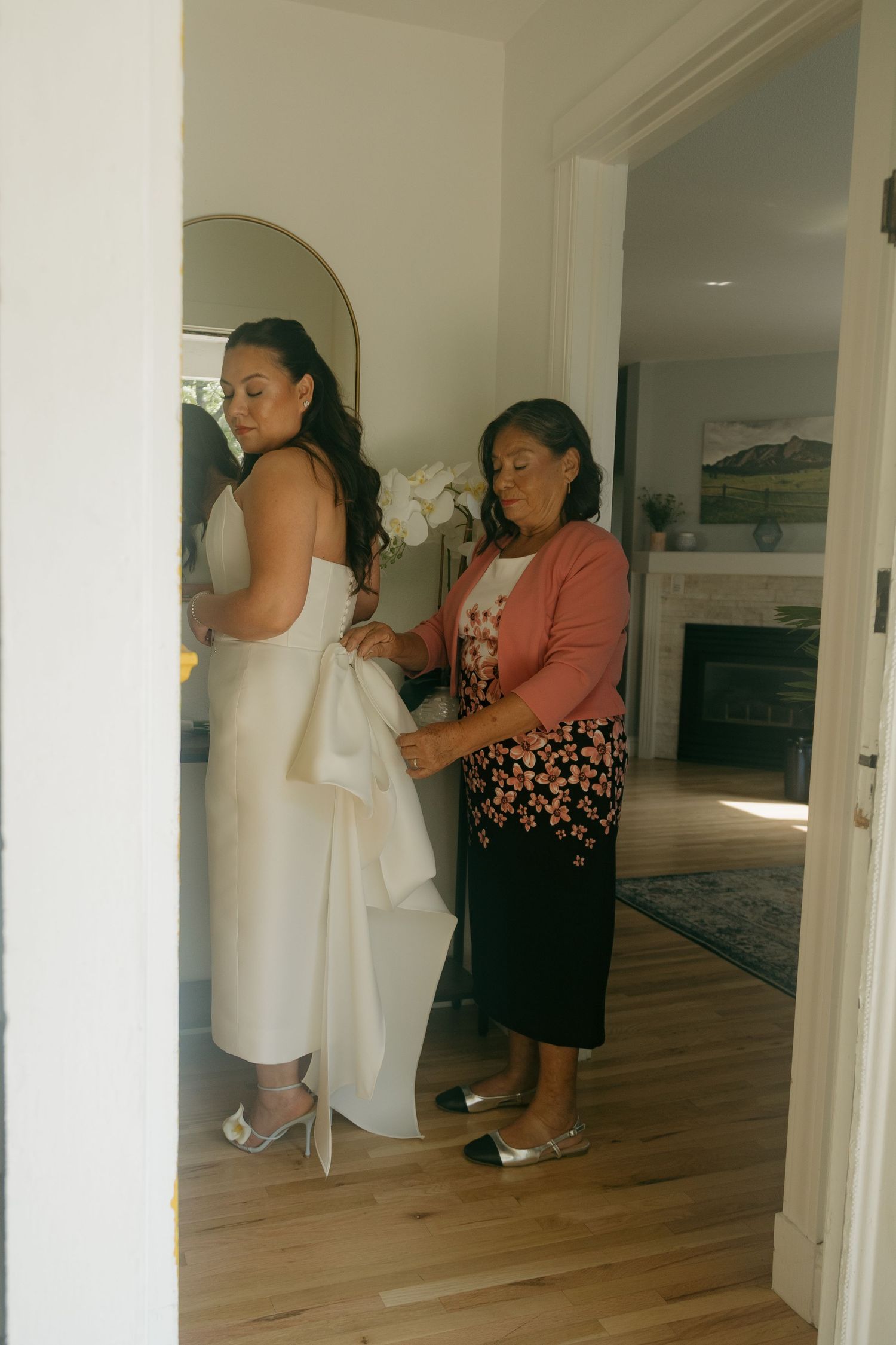 Mother helps bride into white wedding gown in a sunlit room with hardwood floors.