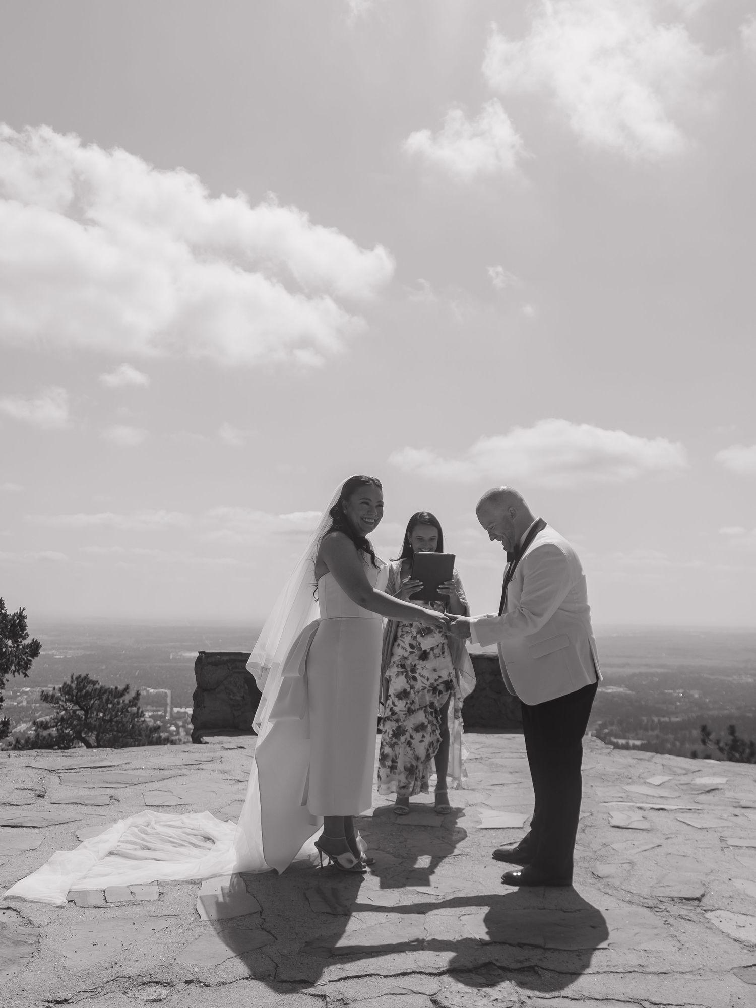 Black and white photo of a couple exchanging vows with an officiant on a scenic mountain overlook on their wedding day.