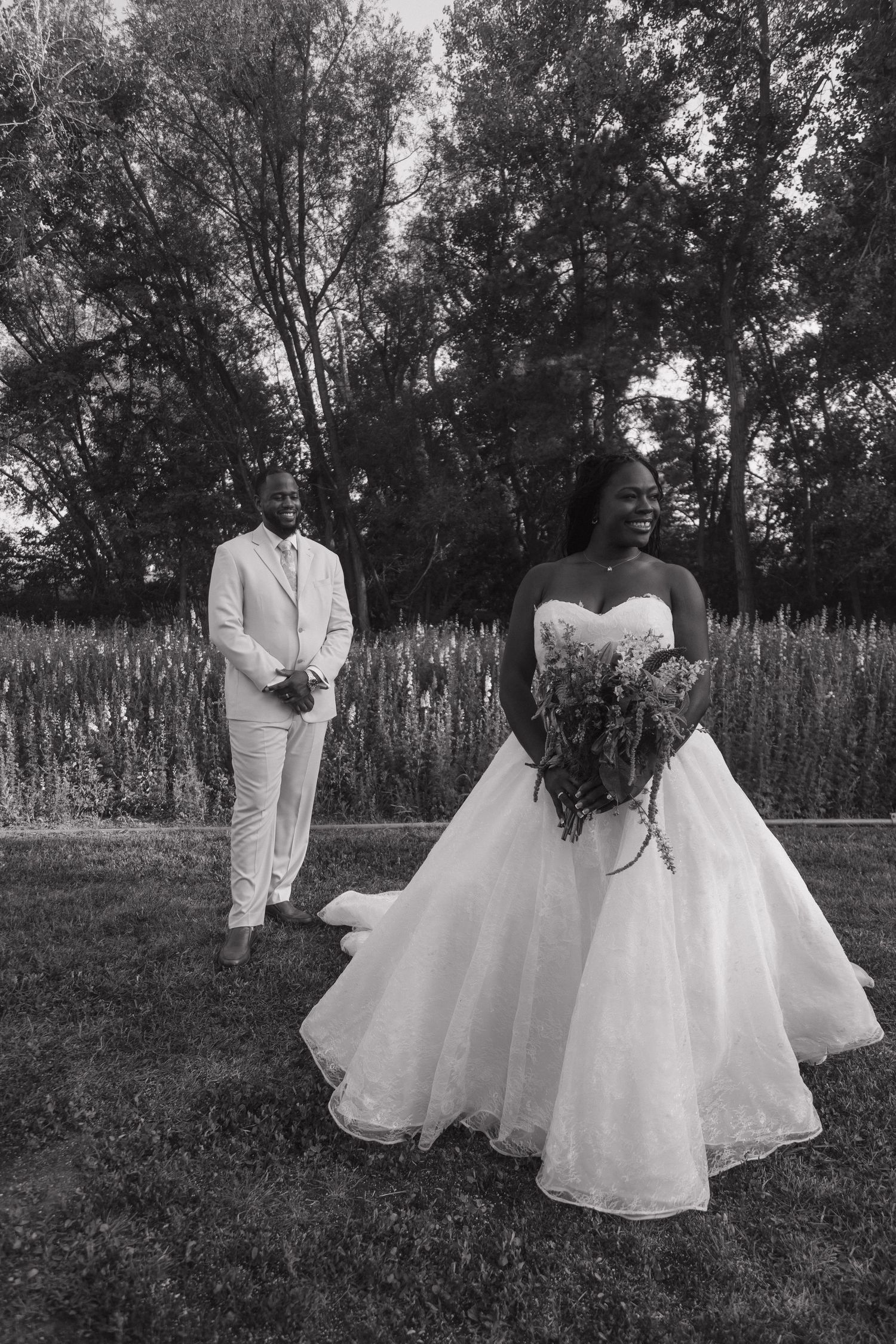 Black and white photo of a bride holding a bouquet in a ball gown as the groom stands behind her outdoors.