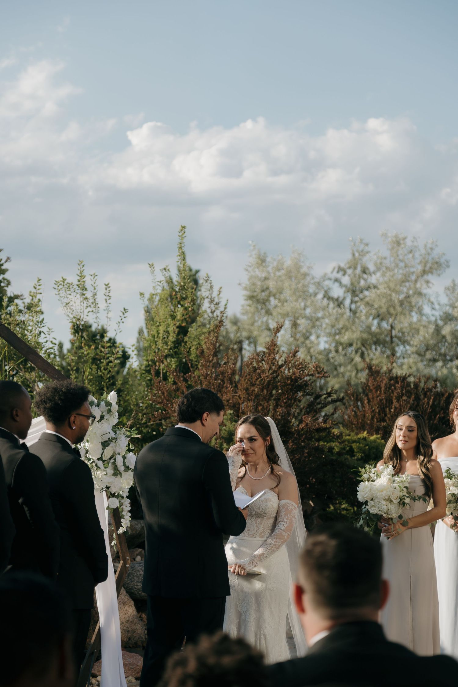 Bride smiling at groom during outdoor wedding ceremony with white floral arch backdrop.