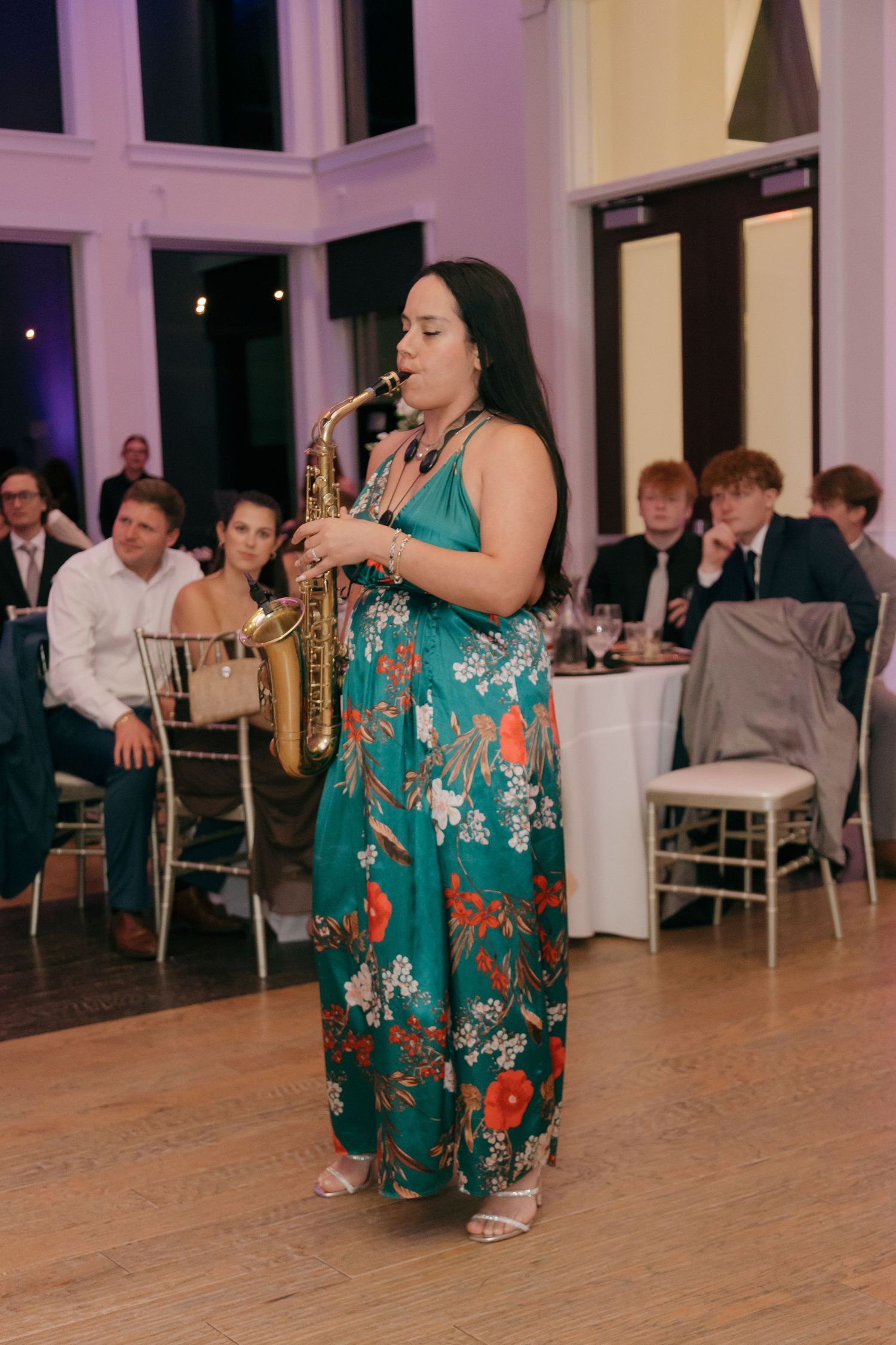 Musician in floral green dress playing saxophone at elegant wedding reception for seated guests.