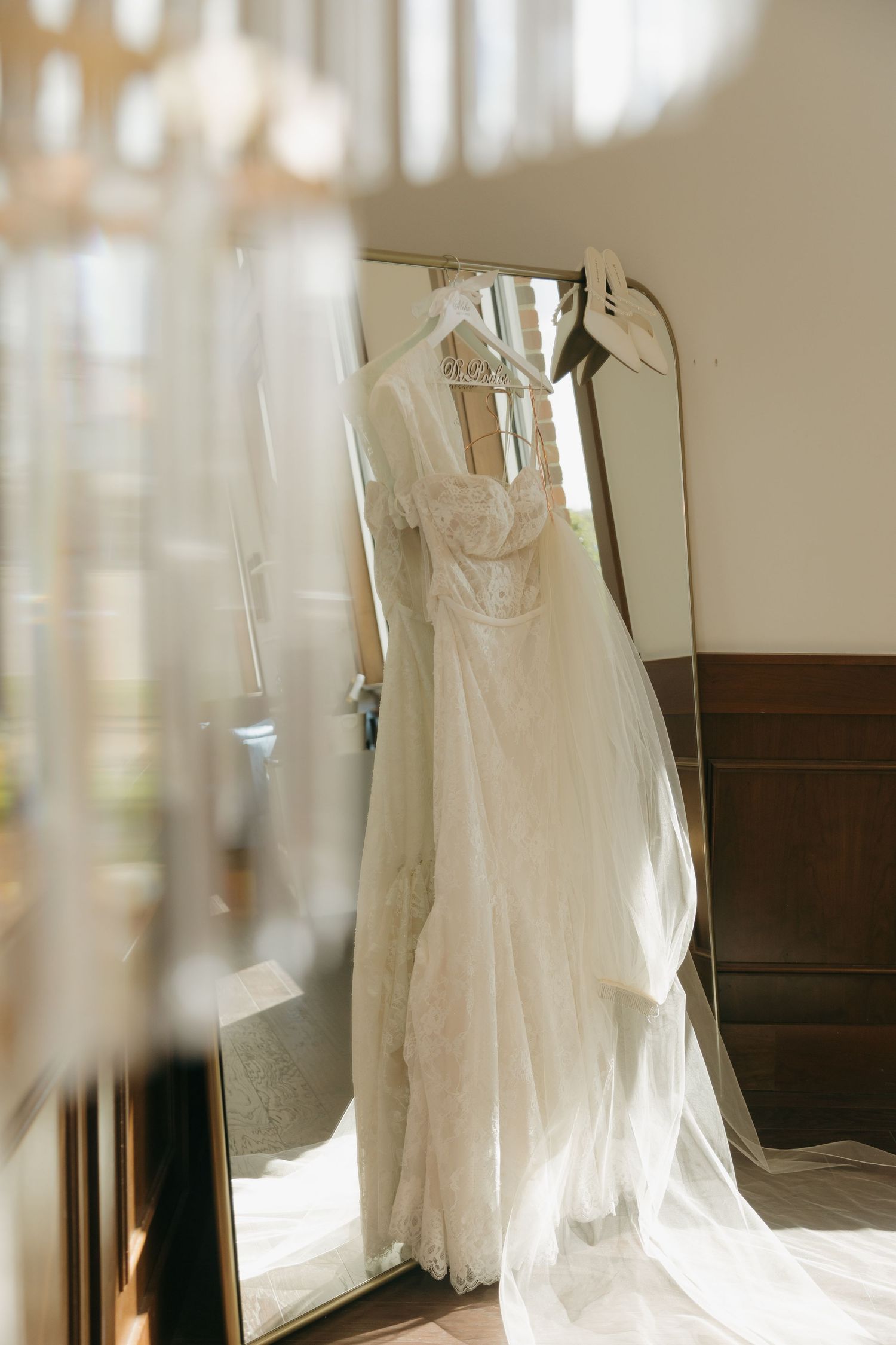 Elegant white lace wedding dress with cap sleeves hanging on a mirror rack in soft natural light.