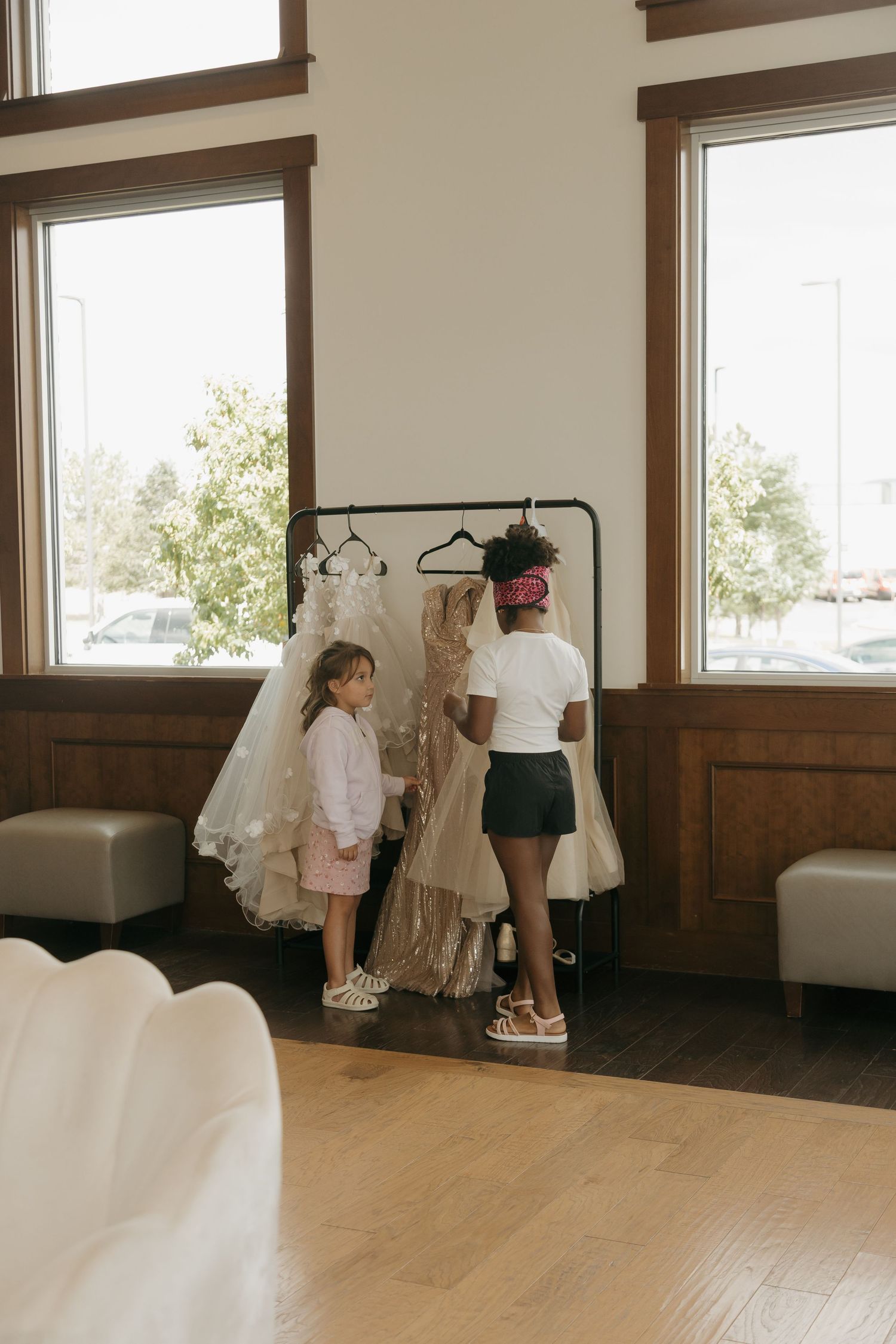 Two young girls in a bridal boutique admiring wedding dresses on a clothing rack near large windows.