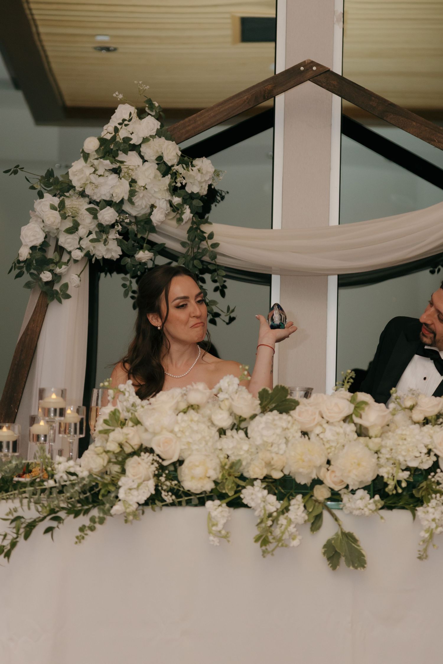Elegant bride sits at floral sweetheart table adorned with white roses and greenery at reception.
