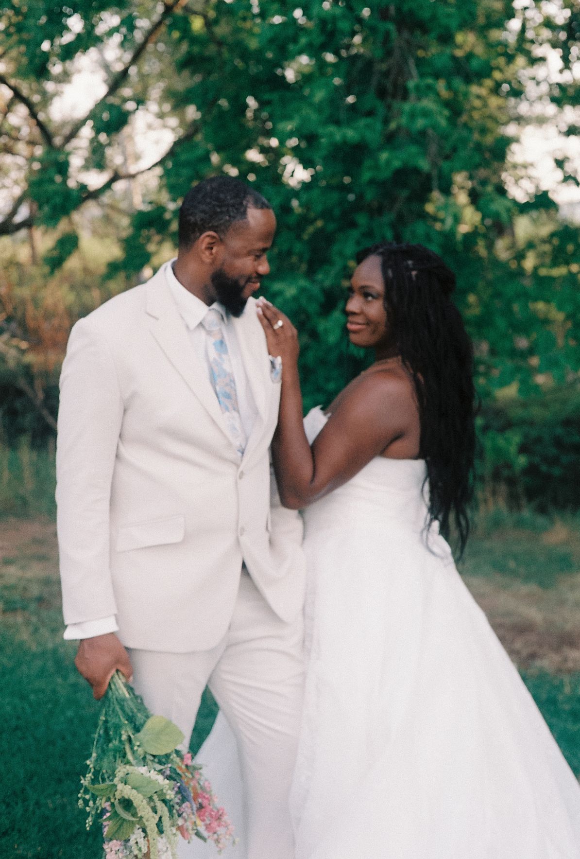 A bride and groom in white wedding attire share a romantic moment outdoors surrounded by lush green trees.