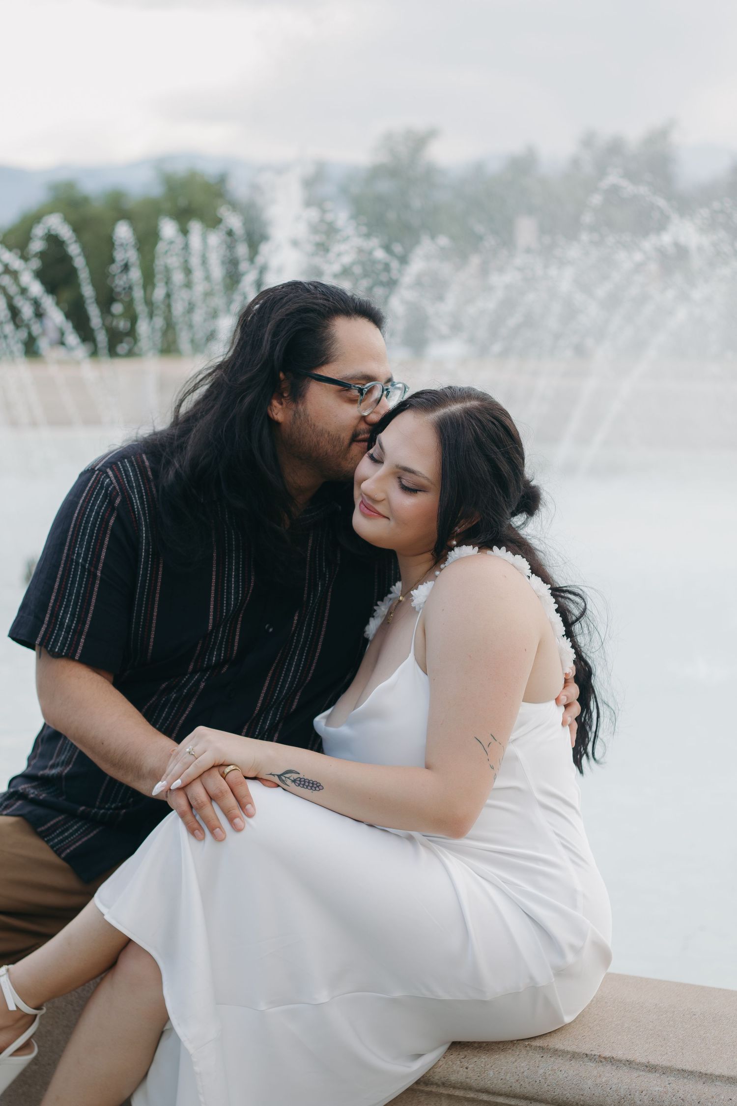 A tender embrace shared between a couple near a fountain with water spraying in the background.