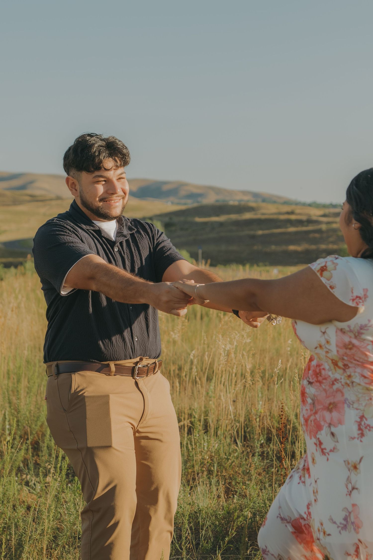 A couple dances playfully in a field with mountains in the background during golden hour.