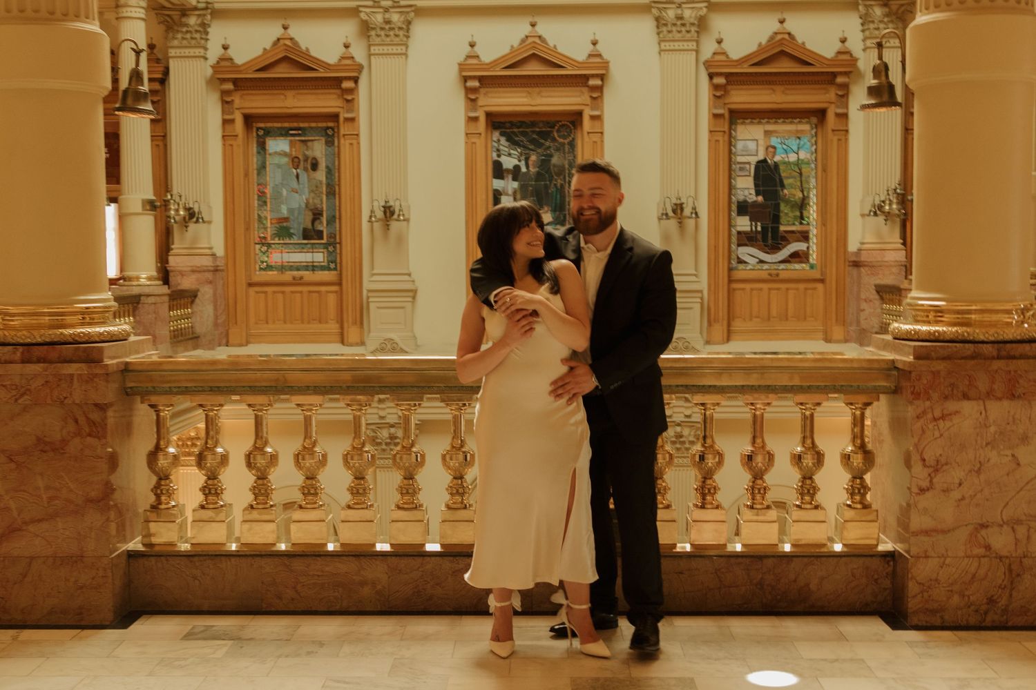 An elegant couple shares an intimate moment on a grand marble balcony surrounded by ornate architectural details and columns.