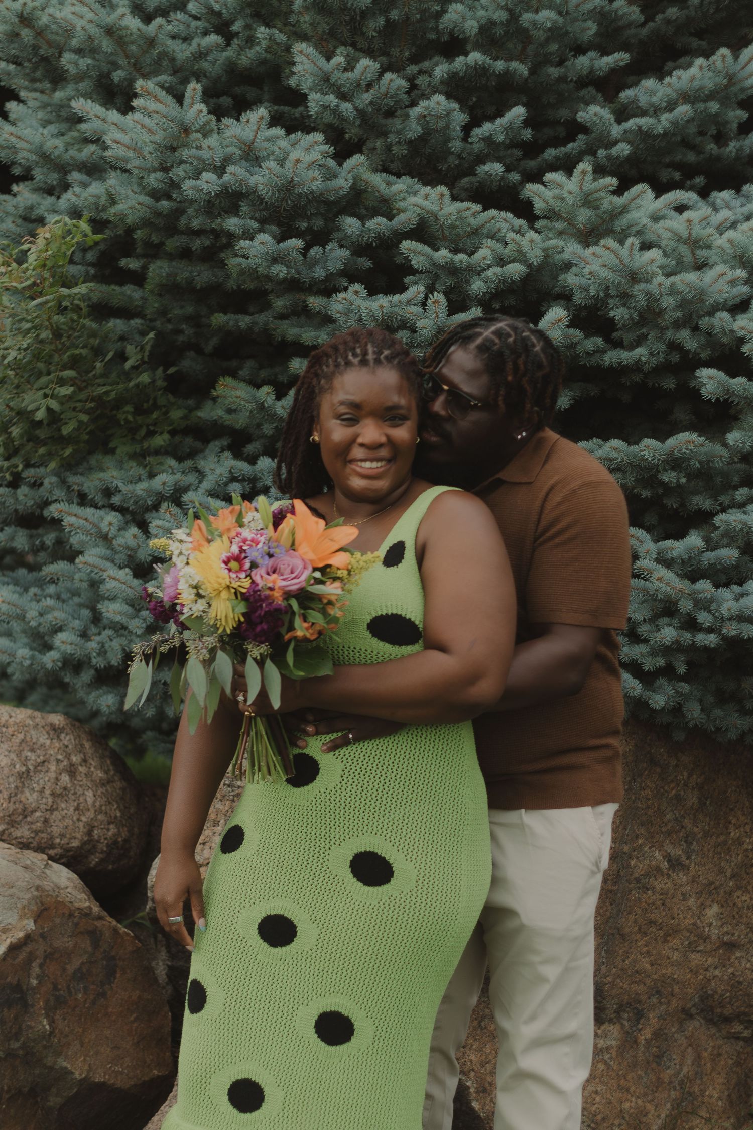 A romantic couple poses in front of blue spruce trees with a colorful bouquet and a green polka dot dress.