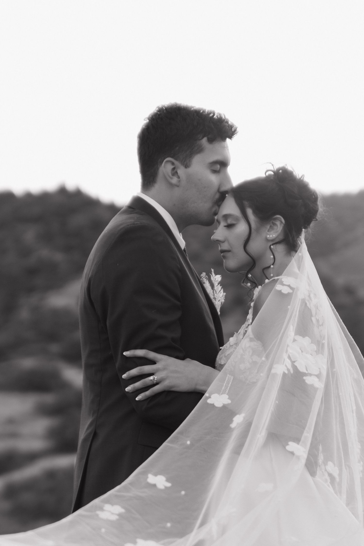 Black and white portrait shows tender moment under flowing bridal veil.