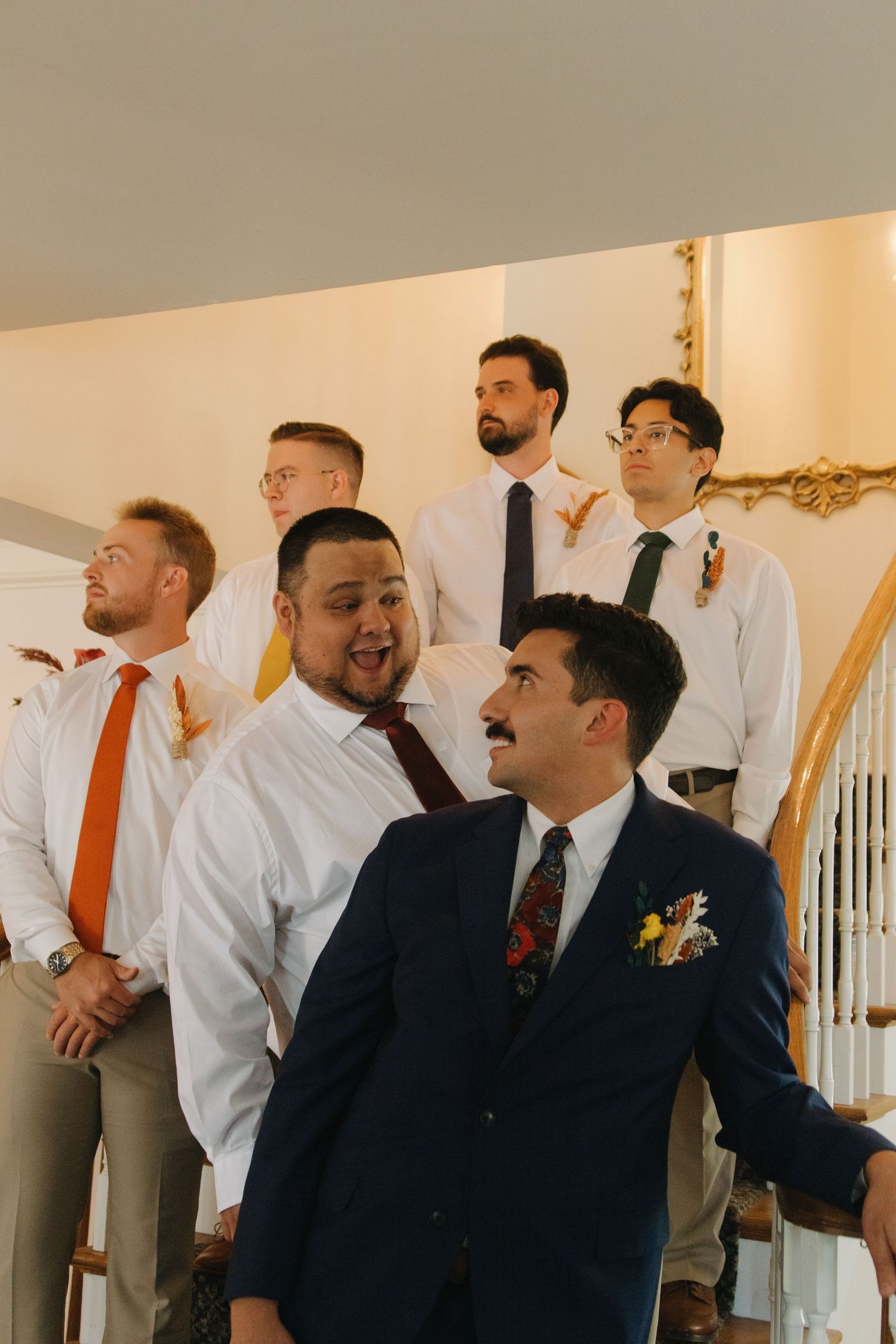 Groomsmen in white shirts with orange ties laugh together during pre-wedding preparations.