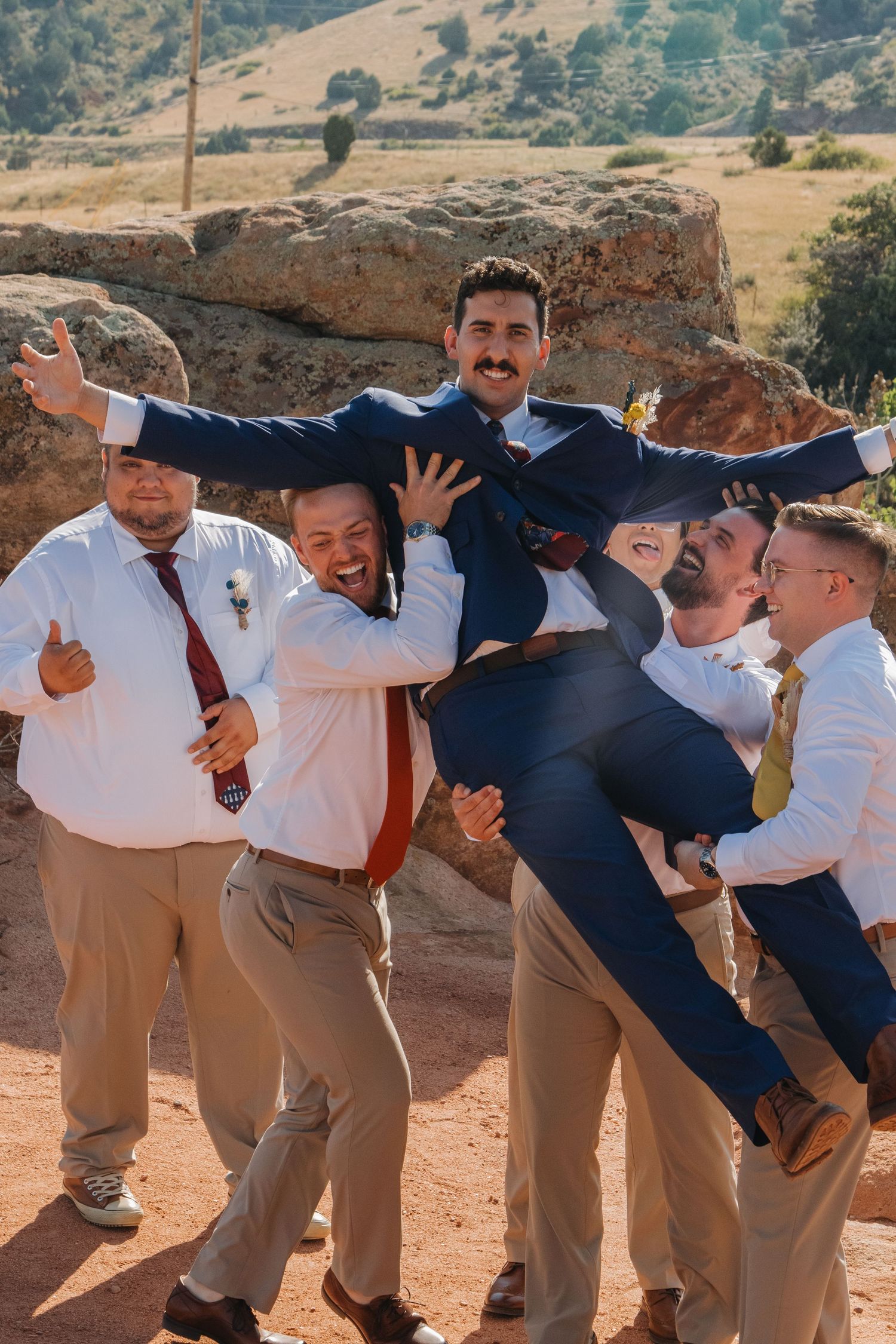 Groomsmen in navy suits and khakis posing playfully on red rock formations.