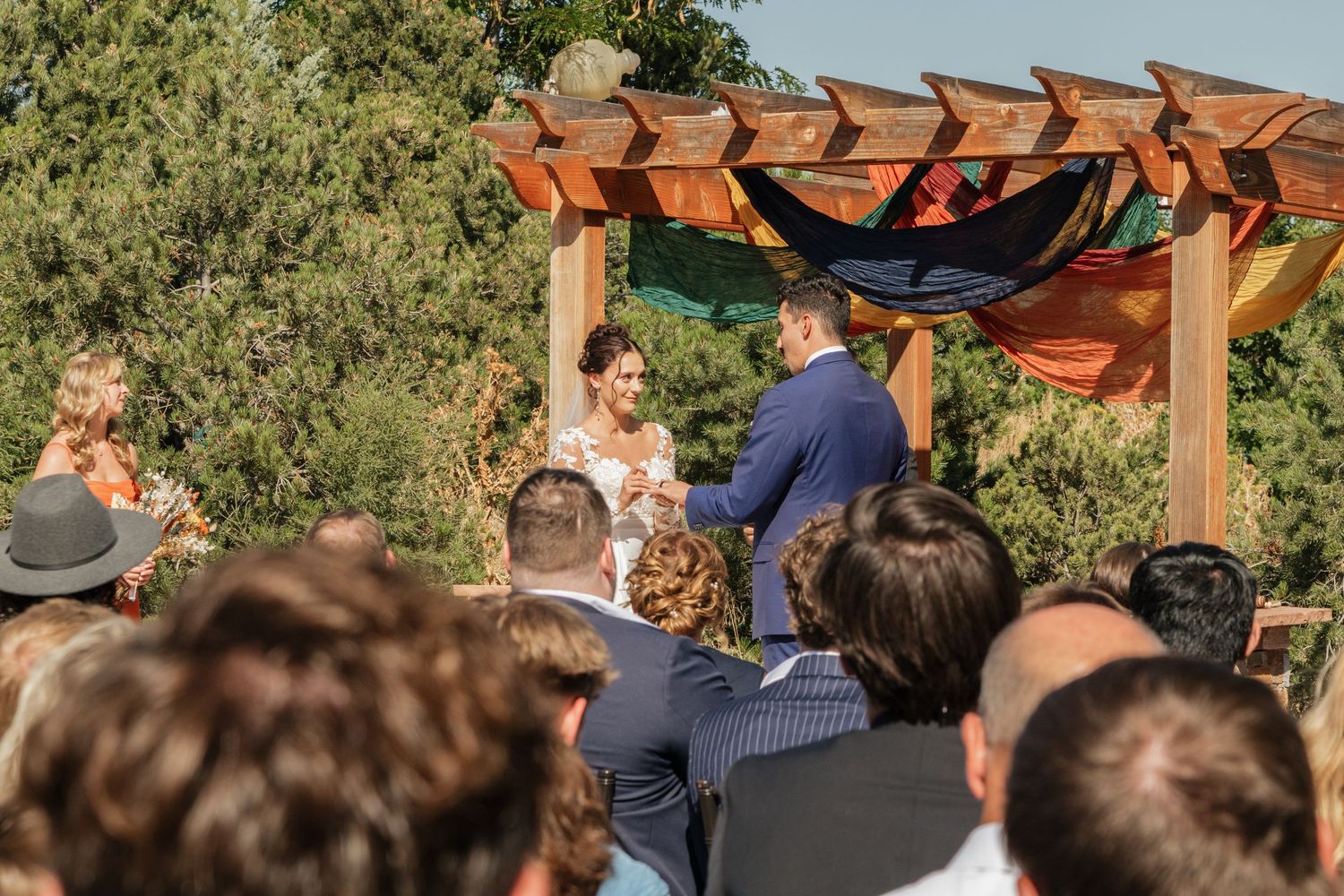 Wedding guests seated in rows watching an outdoor ceremony under a wooden pergola.
