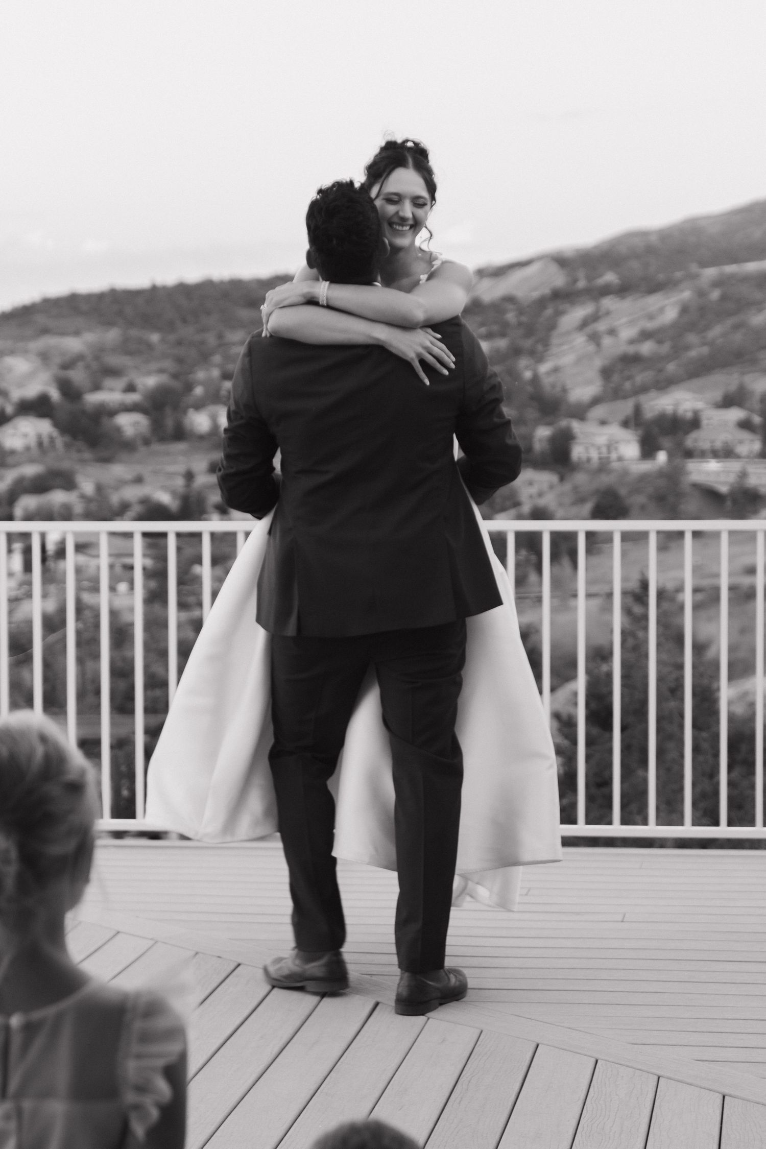 Two figures embrace on a white deck overlooking a mountain landscape.