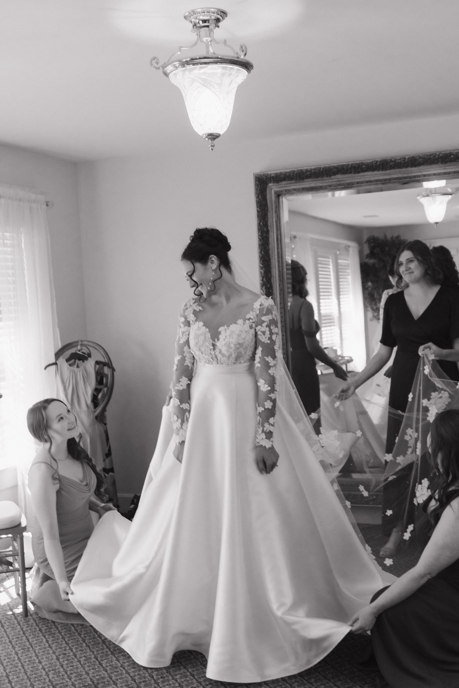 Black and white photo of bridesmaids helping with a wedding dress in a brightly lit room.