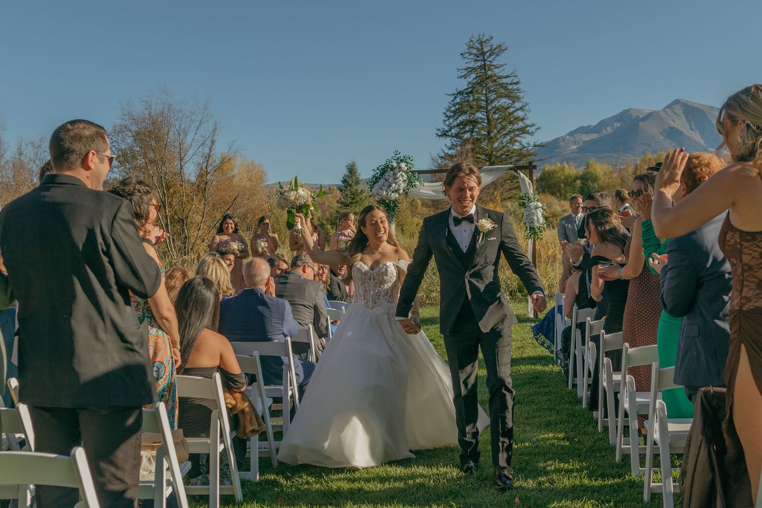 Wedding ceremony guests seated on white chairs on a green lawn with mountain views in the background during sunset.