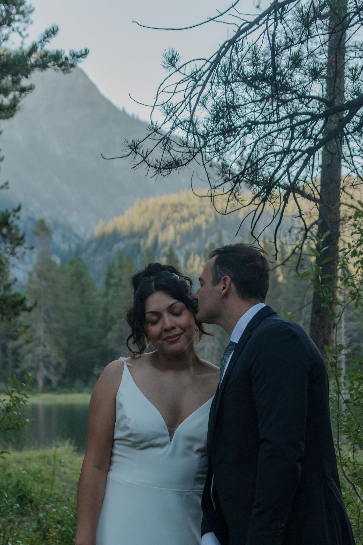 A tender wedding moment captured against evergreen trees and mountain scenery.