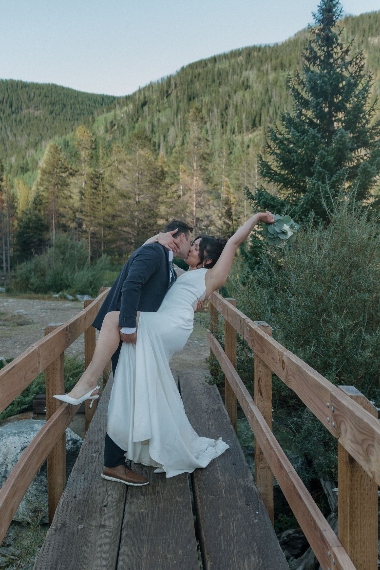 A couple shares a romantic dip pose on a wooden bridge surrounded by evergreen forest scenery.