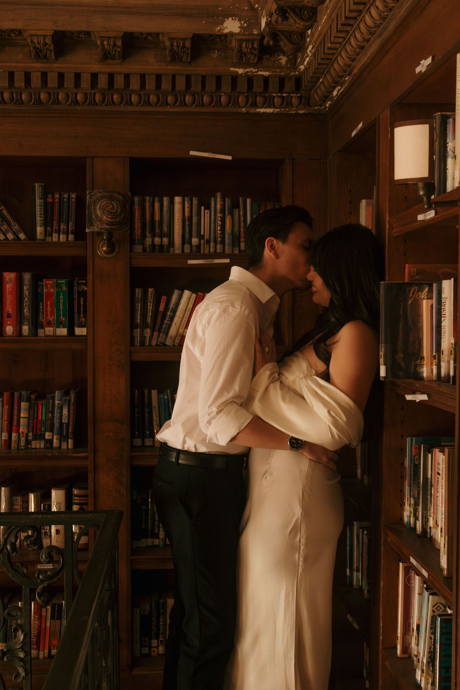 A couple shares an intimate moment in a library surrounded by wooden bookshelves.