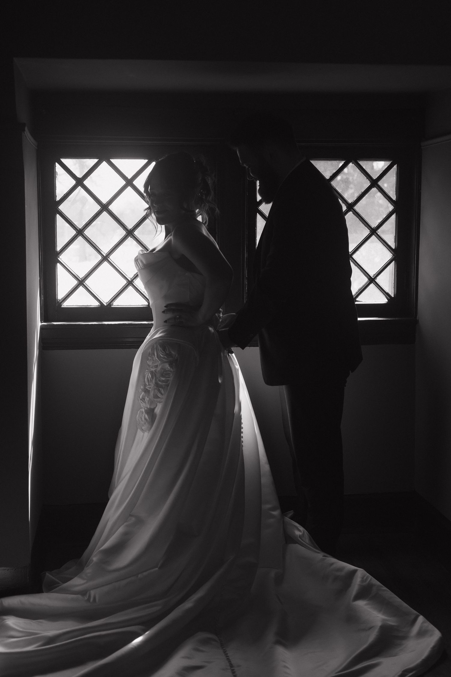 Silhouettes of a couple in formal wear standing by latticed windows in dramatic lighting.